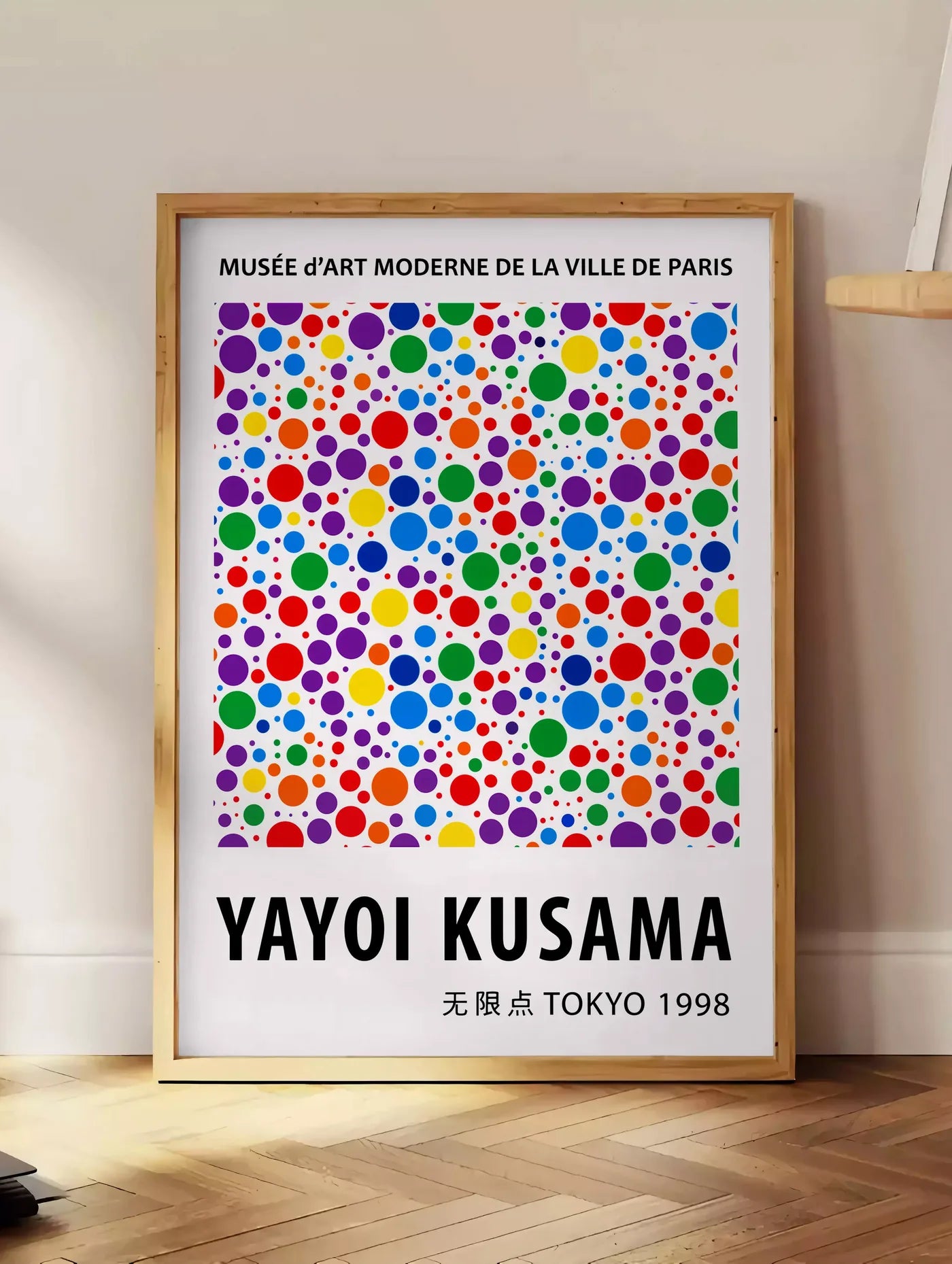 Estampe affiche pois multicolores de Kusama art japonais, posee au sol dans un coin salon, sur un parquet a chevrons, pres d'une table d'appoint en chene chargee de livres