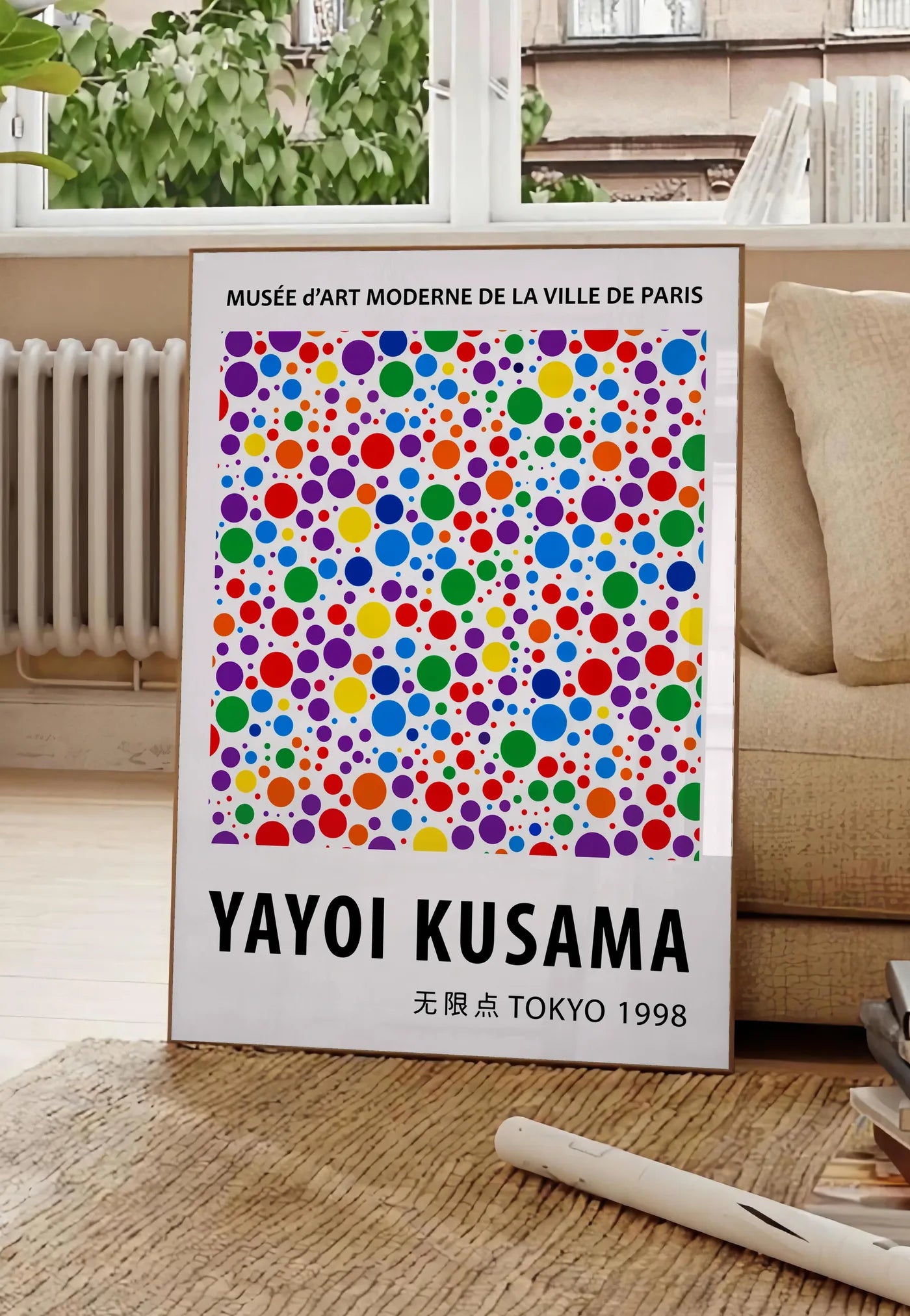 Poster affiche pois multicolores de Kusama art japonais, posee au sol dans un salon parisien, devant une grande fenetre donnant sur un immeuble haussmannien, entre un ficus et un canape creme