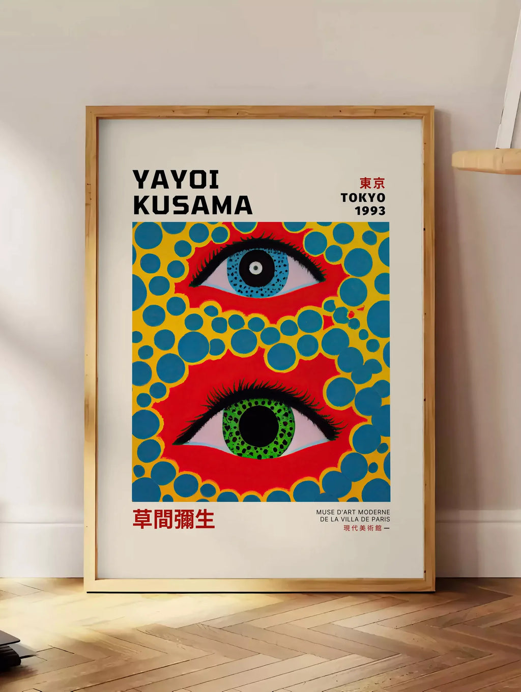 Estampe affiche yeux a pois jaune de Kusama art japonais, posee au sol dans un coin salon, sur un parquet a chevrons, pres d'une table d'appoint en chene chargee de livres