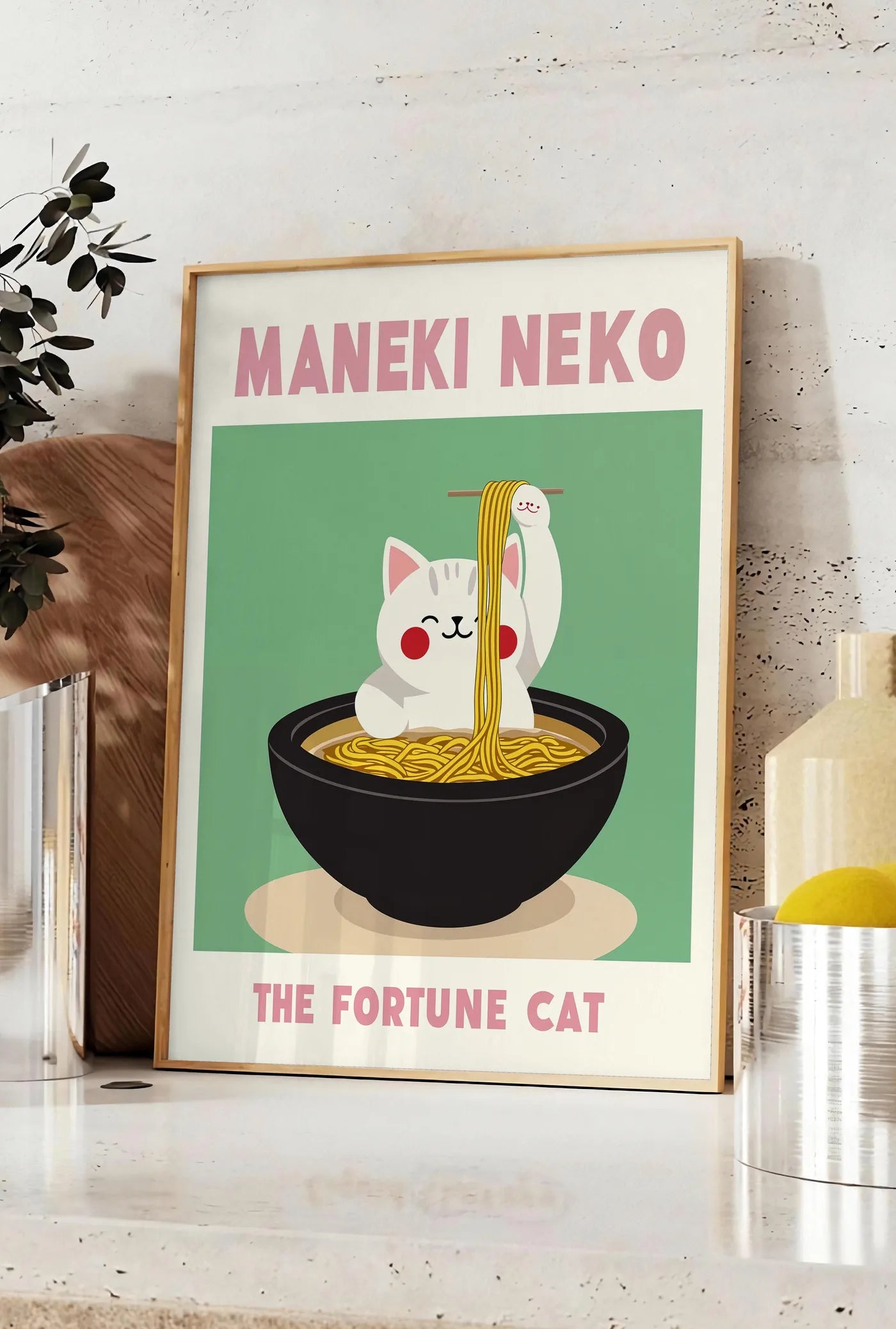 Affiche affiche Maneki Neko chat et ramen sur fond vert sauge, posee sur un plan de travail de cuisine en pierre claire, entre une branche d'eucalyptus en vase et un bocal de citrons