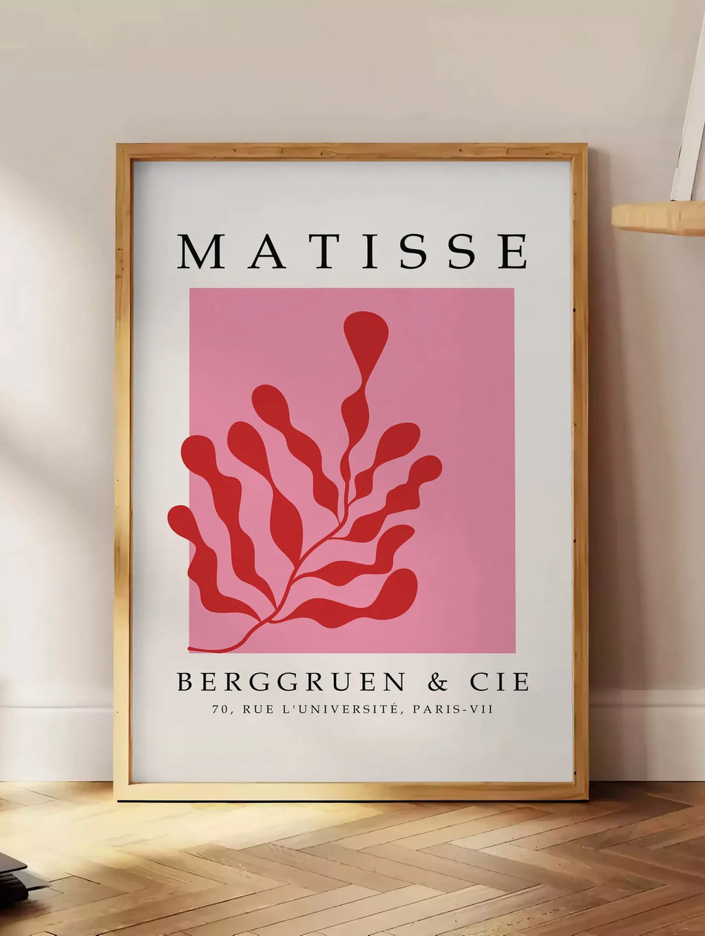 Estampe algue Rouge sur Rose en papiers decoupes Matisse sur fond colore, posee au sol dans un coin salon, sur un parquet a chevrons, pres d'une table d'appoint en chene chargee de livres