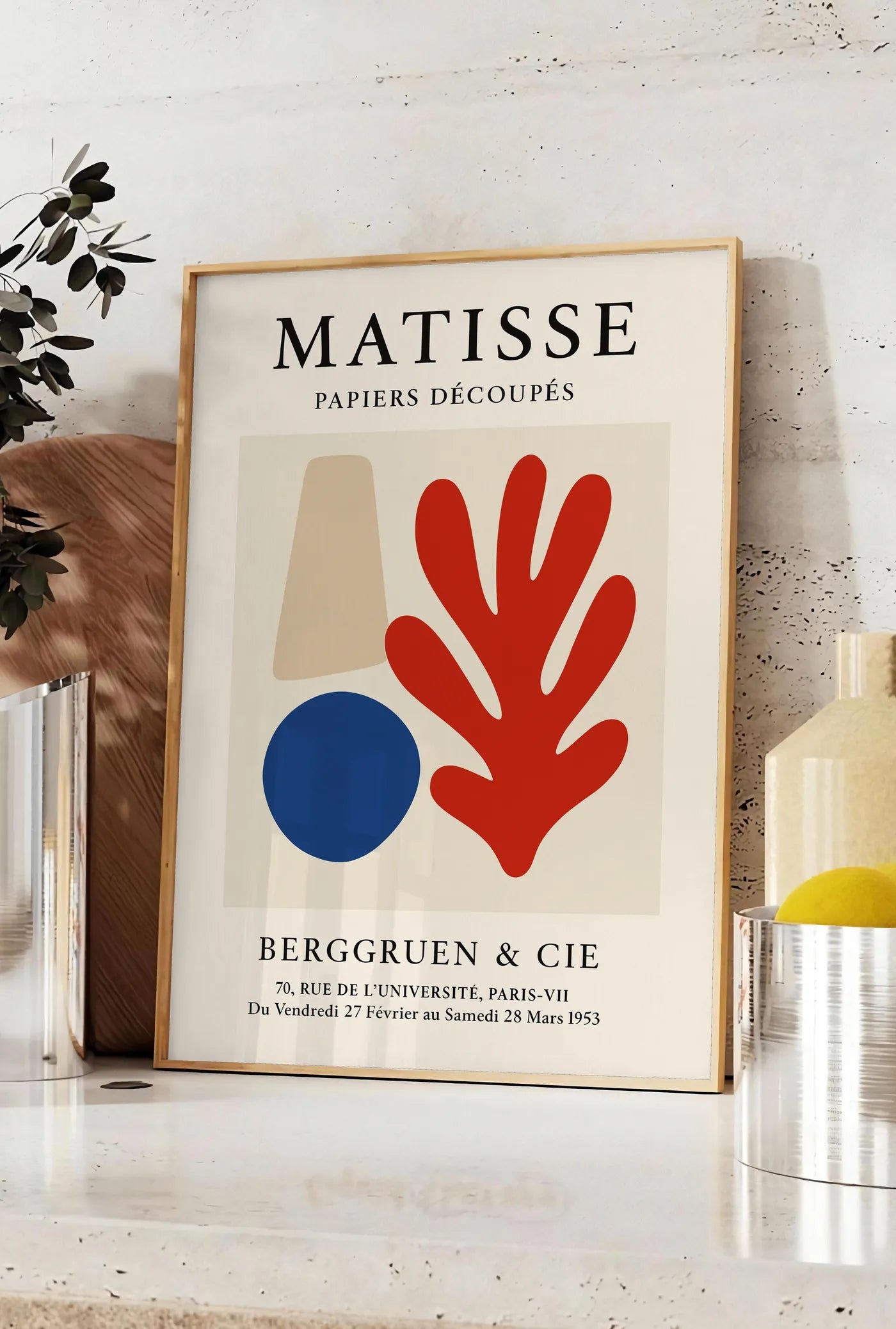 Affiche cone beige cercle bleu et corail rouge sur fond creme, posee sur un plan de travail de cuisine en pierre claire, entre une branche d'eucalyptus en vase et un bocal de citrons