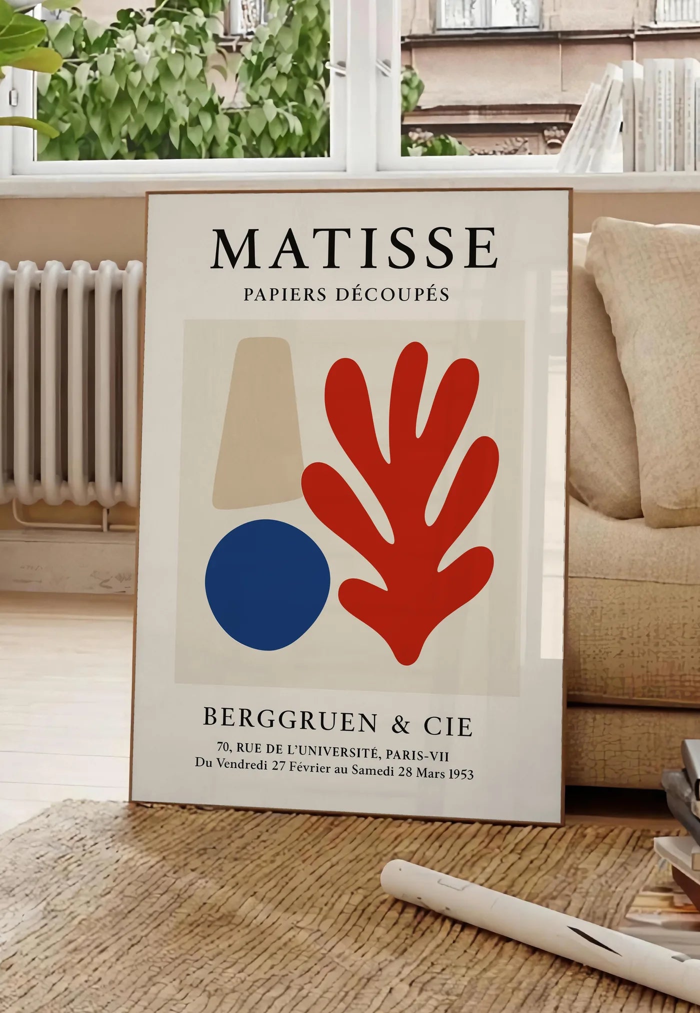 Poster cone beige cercle bleu et corail rouge sur fond creme, posee au sol dans un salon parisien, devant une grande fenetre donnant sur un immeuble haussmannien, entre un ficus et un canape creme
