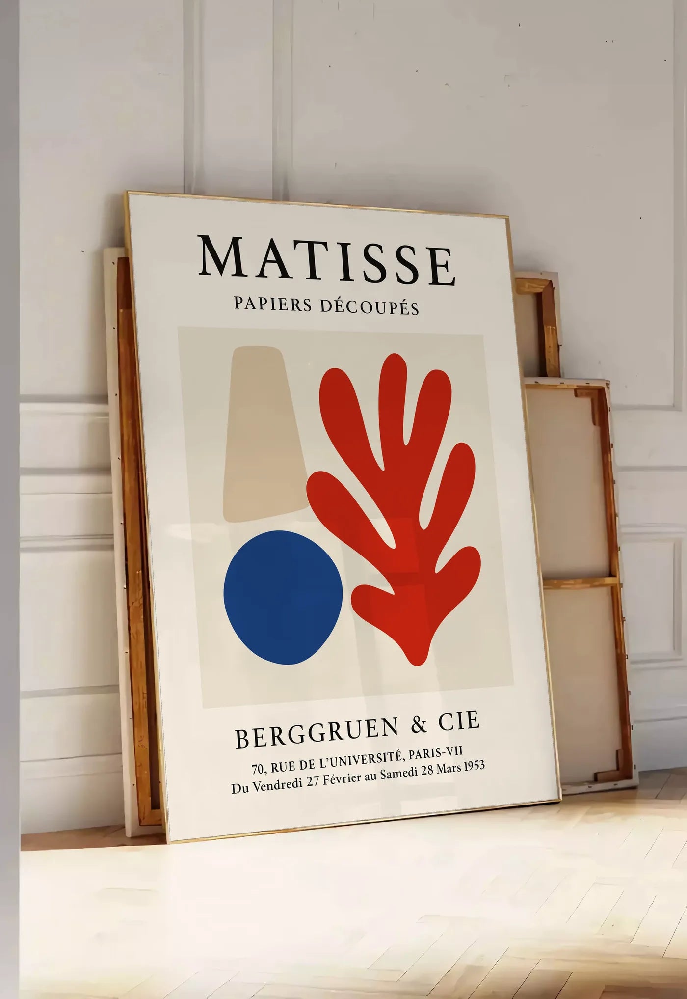 Tirage encadrable cone beige cercle bleu et corail rouge sur fond creme, posee au sol dans une entree lumineuse, contre un mur a panneaux blancs, a cote de chassis en bois empiles