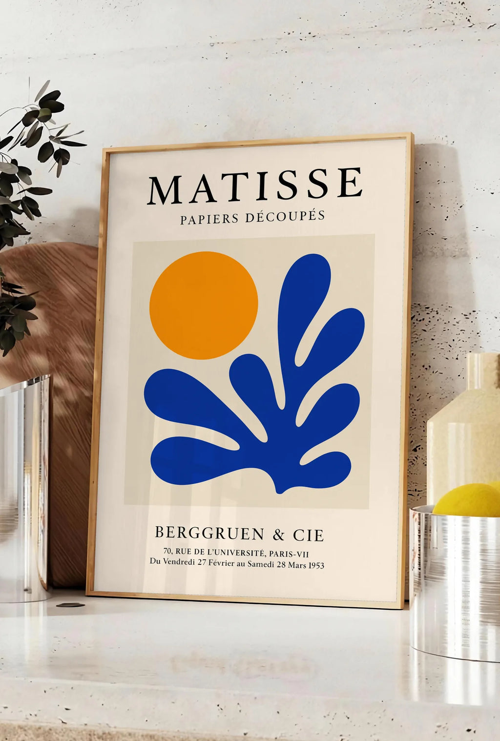 Affiche corail Bleu et Soleil Orange en papiers decoupes Matisse sur fond colore, posee sur un plan de travail de cuisine en pierre claire, entre une branche d'eucalyptus en vase et un bocal de citrons