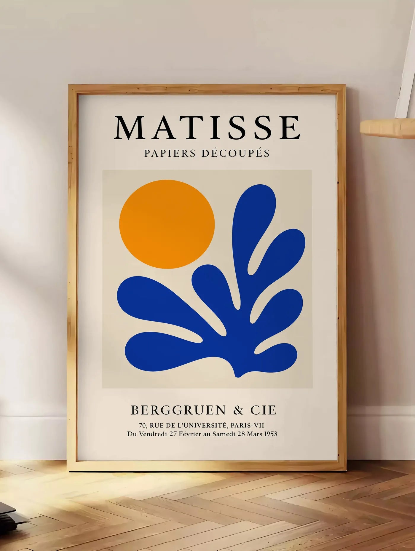 Estampe corail Bleu et Soleil Orange en papiers decoupes Matisse sur fond colore, posee au sol dans un coin salon, sur un parquet a chevrons, pres d'une table d'appoint en chene chargee de livres