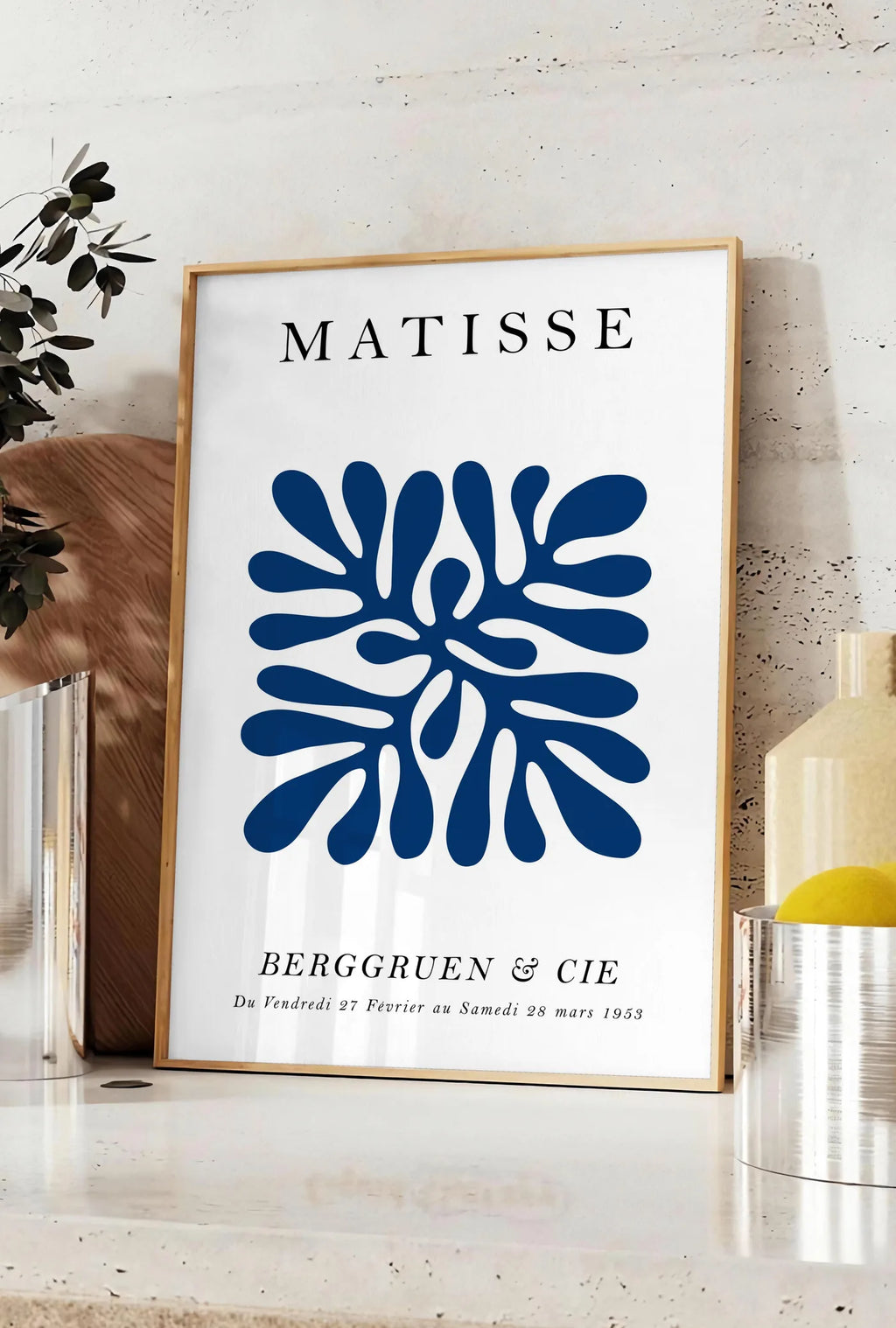 Affiche corail Bleu Symetrique en papiers decoupes Matisse sur fond colore, posee sur un plan de travail de cuisine en pierre claire, entre une branche d'eucalyptus en vase et un bocal de citrons