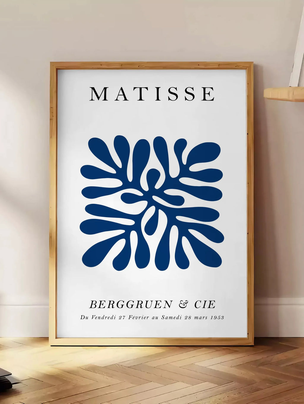 Estampe corail Bleu Symetrique en papiers decoupes Matisse sur fond colore, posee au sol dans un coin salon, sur un parquet a chevrons, pres d'une table d'appoint en chene chargee de livres