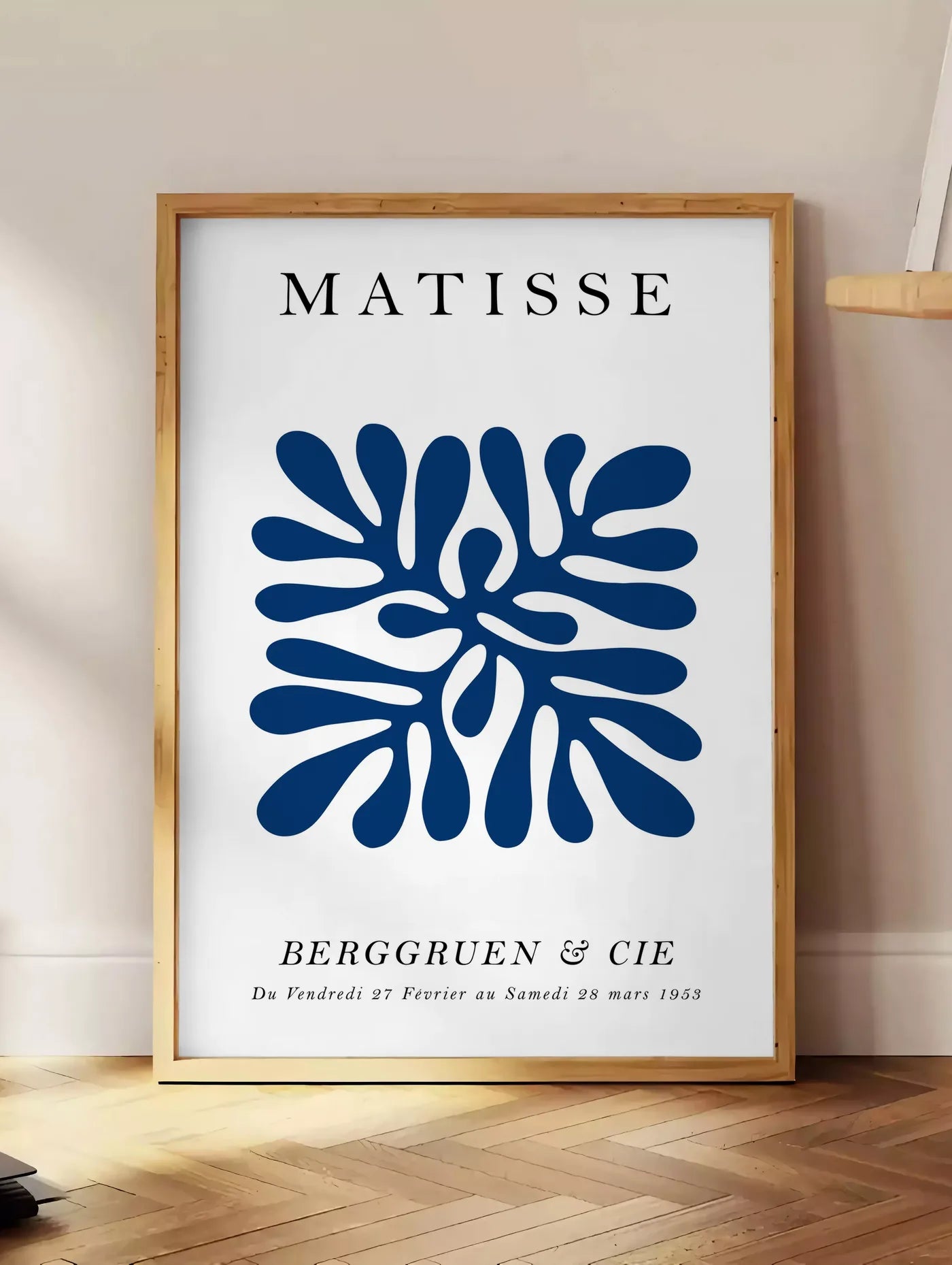 Estampe corail Bleu Symetrique en papiers decoupes Matisse sur fond colore, posee au sol dans un coin salon, sur un parquet a chevrons, pres d'une table d'appoint en chene chargee de livres