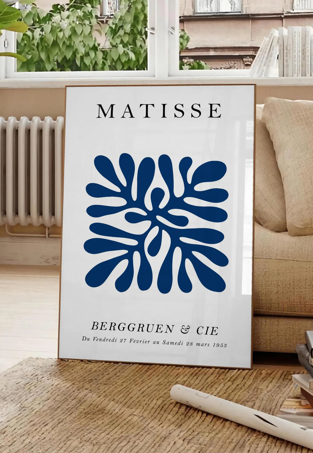 Poster corail Bleu Symetrique en papiers decoupes Matisse sur fond colore, posee au sol dans un salon parisien, devant une grande fenetre donnant sur un immeuble haussmannien, entre un ficus et un canape creme