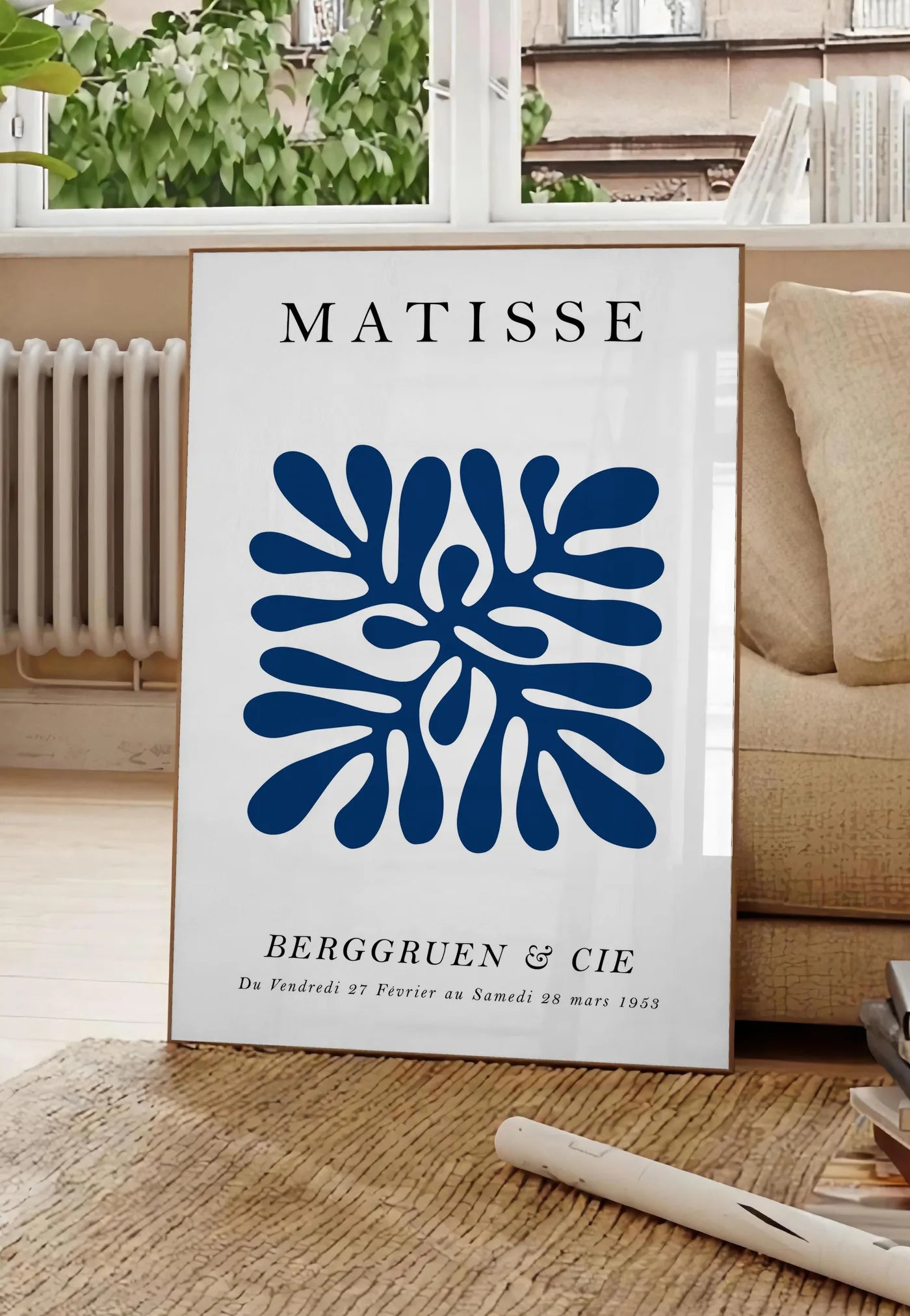 Poster corail Bleu Symetrique en papiers decoupes Matisse sur fond colore, posee au sol dans un salon parisien, devant une grande fenetre donnant sur un immeuble haussmannien, entre un ficus et un canape creme