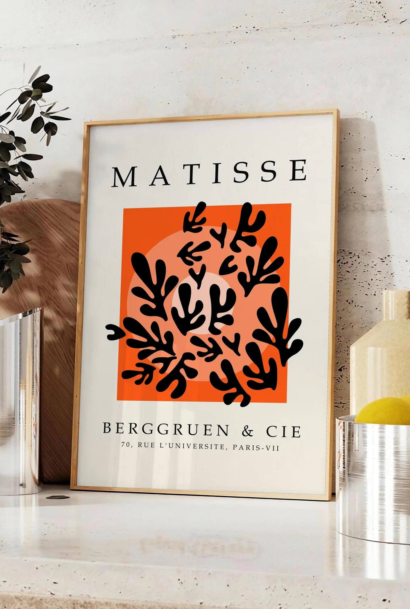 Affiche corail Noir sur Orange en papiers decoupes Matisse sur fond colore, posee sur un plan de travail de cuisine en pierre claire, entre une branche d'eucalyptus en vase et un bocal de citrons