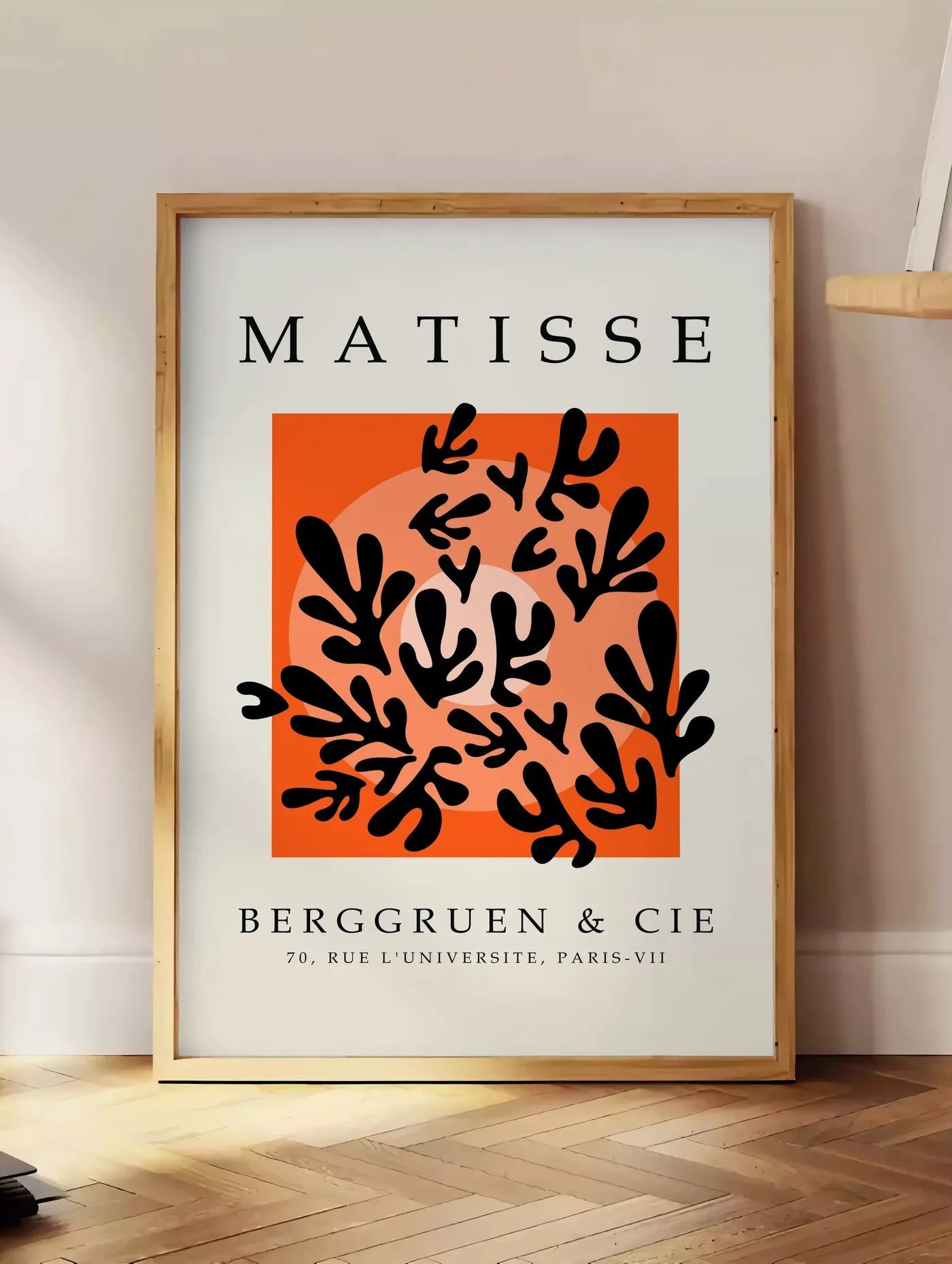 Estampe corail Noir sur Orange en papiers decoupes Matisse sur fond colore, posee au sol dans un coin salon, sur un parquet a chevrons, pres d'une table d'appoint en chene chargee de livres