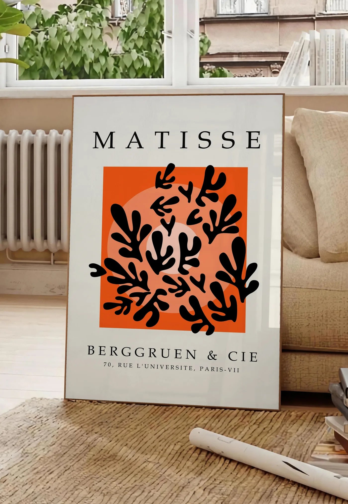 Poster corail Noir sur Orange en papiers decoupes Matisse sur fond colore, posee au sol dans un salon parisien, devant une grande fenetre donnant sur un immeuble haussmannien, entre un ficus et un canape creme