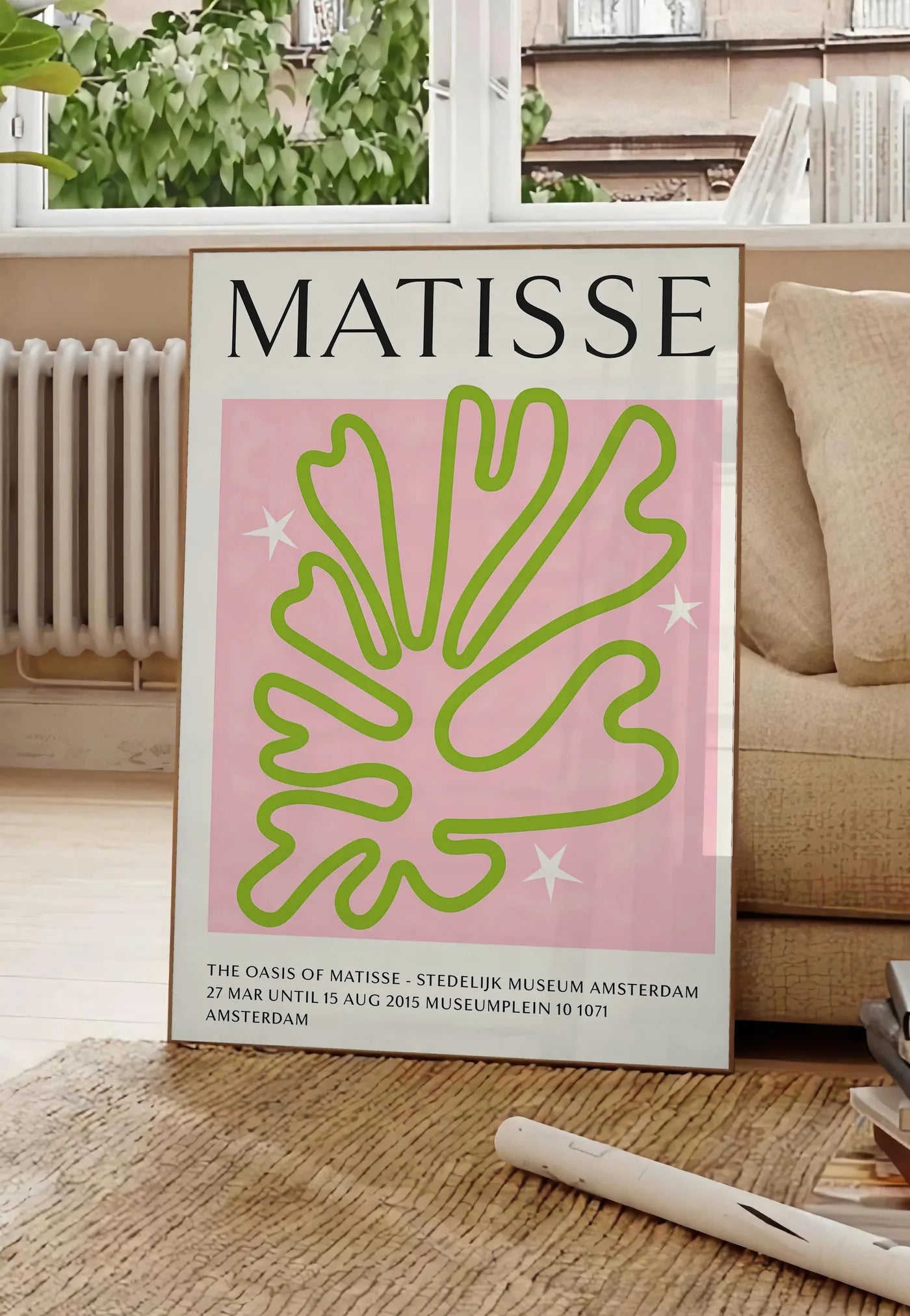 Poster corail Vert Oasis en papiers decoupes Matisse sur fond colore, posee au sol dans un salon parisien, devant une grande fenetre donnant sur un immeuble haussmannien, entre un ficus et un canape creme