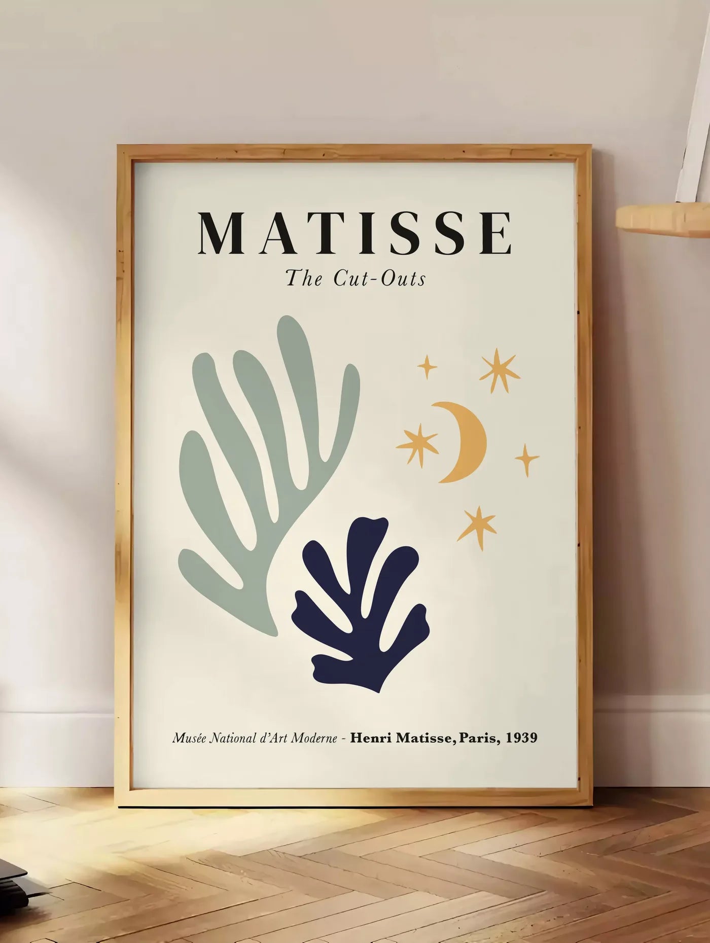 Estampe corail Lune et Etoiles en papiers decoupes Matisse sur fond colore, posee au sol dans un coin salon, sur un parquet a chevrons, pres d'une table d'appoint en chene chargee de livres