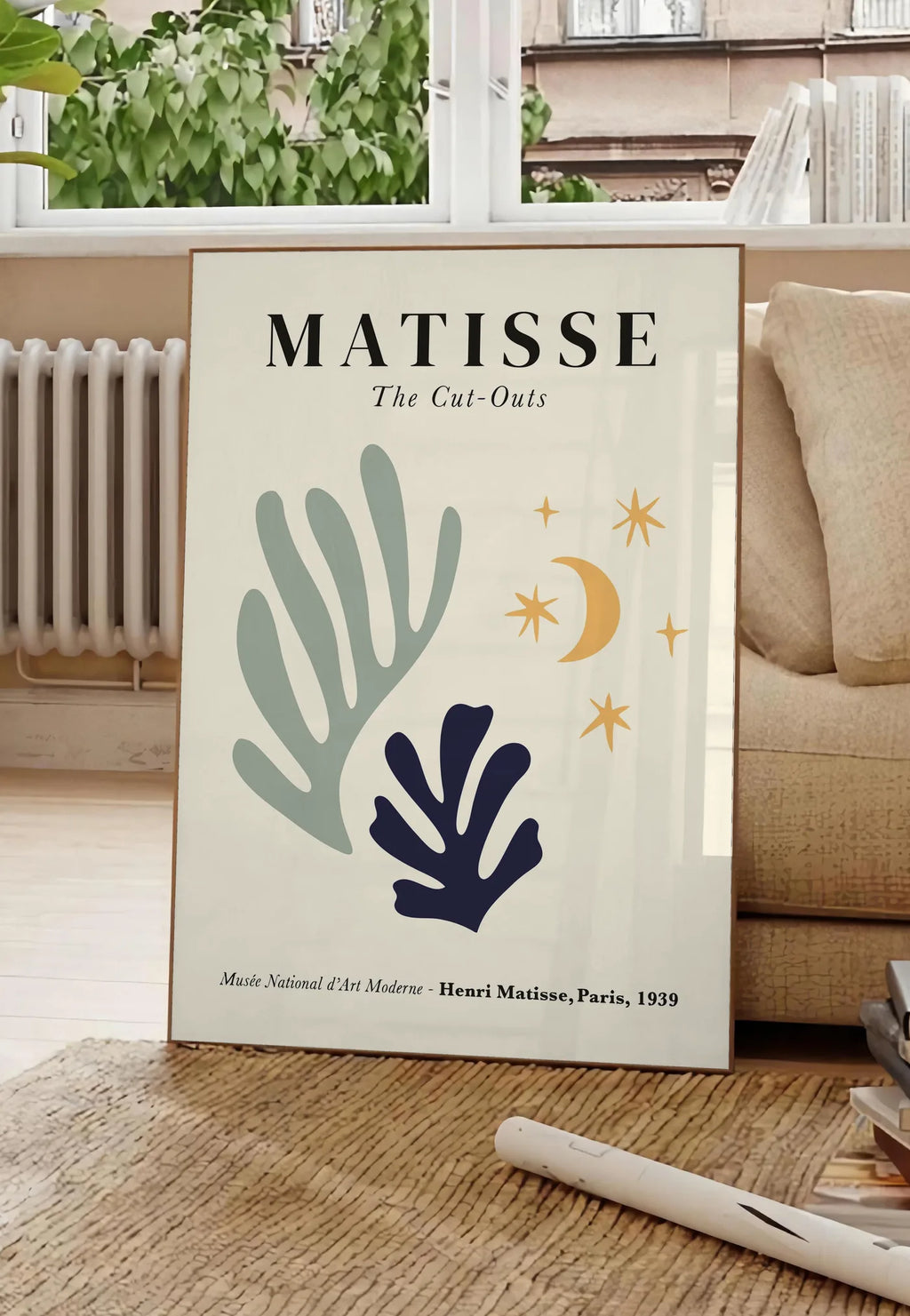 Poster corail Lune et Etoiles en papiers decoupes Matisse sur fond colore, posee au sol dans un salon parisien, devant une grande fenetre donnant sur un immeuble haussmannien, entre un ficus et un canape creme
