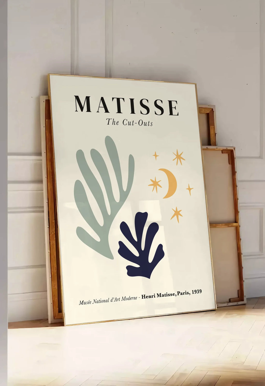 Tirage encadrable corail Lune et Etoiles en papiers decoupes Matisse sur fond colore, posee au sol dans une entree lumineuse, contre un mur a panneaux blancs, a cote de chassis en bois empiles