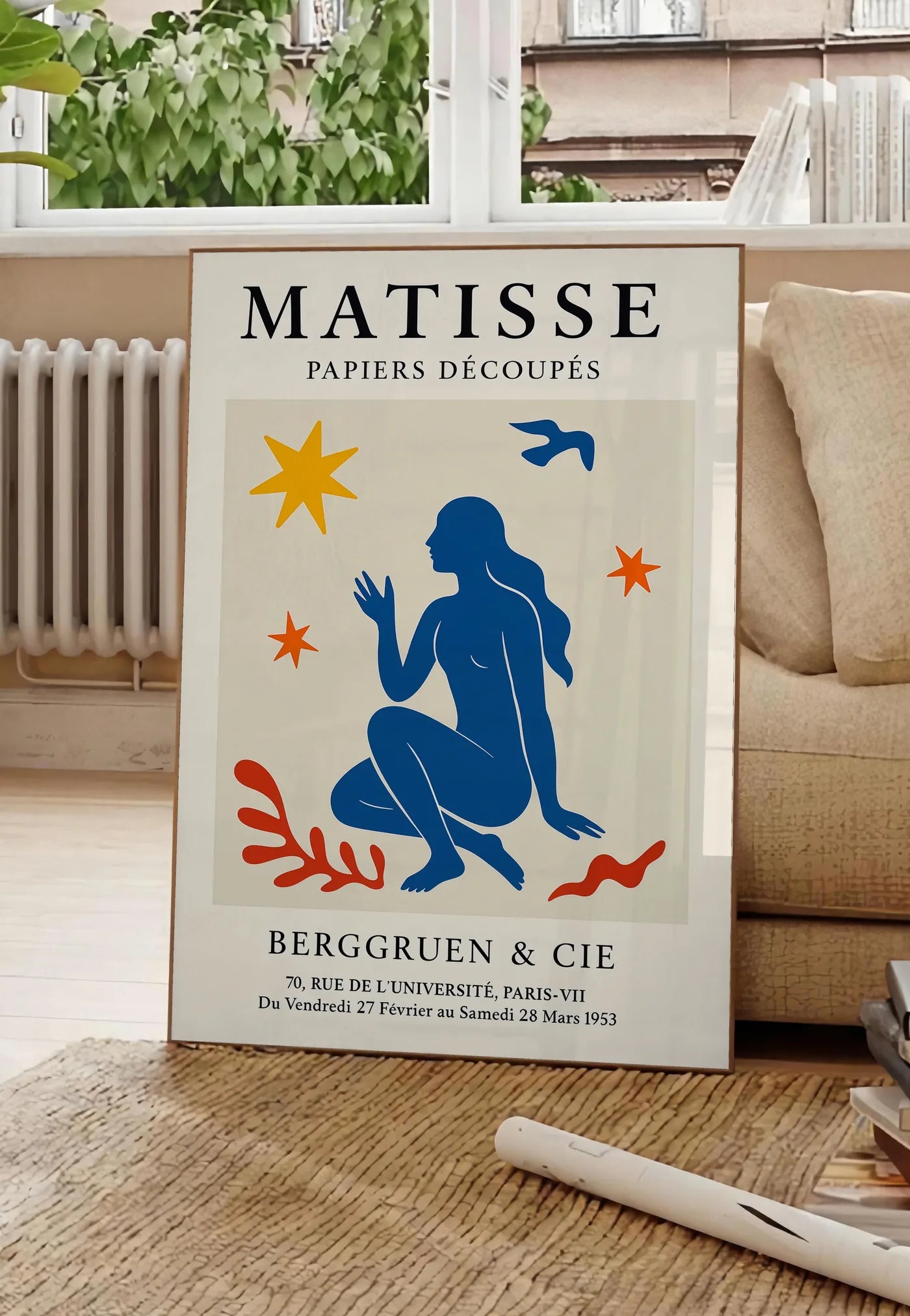 Poster femme bleue parmi etoiles et un oiseau, posee au sol dans un salon parisien, devant une grande fenetre donnant sur un immeuble haussmannien, entre un ficus et un canape creme