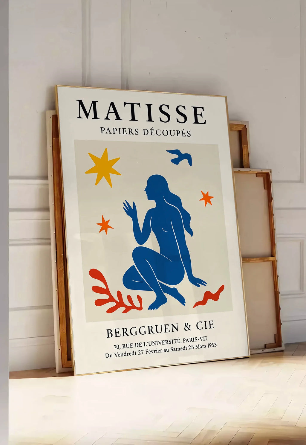Tirage encadrable femme bleue parmi etoiles et un oiseau, posee au sol dans une entree lumineuse, contre un mur a panneaux blancs, a cote de chassis en bois empiles