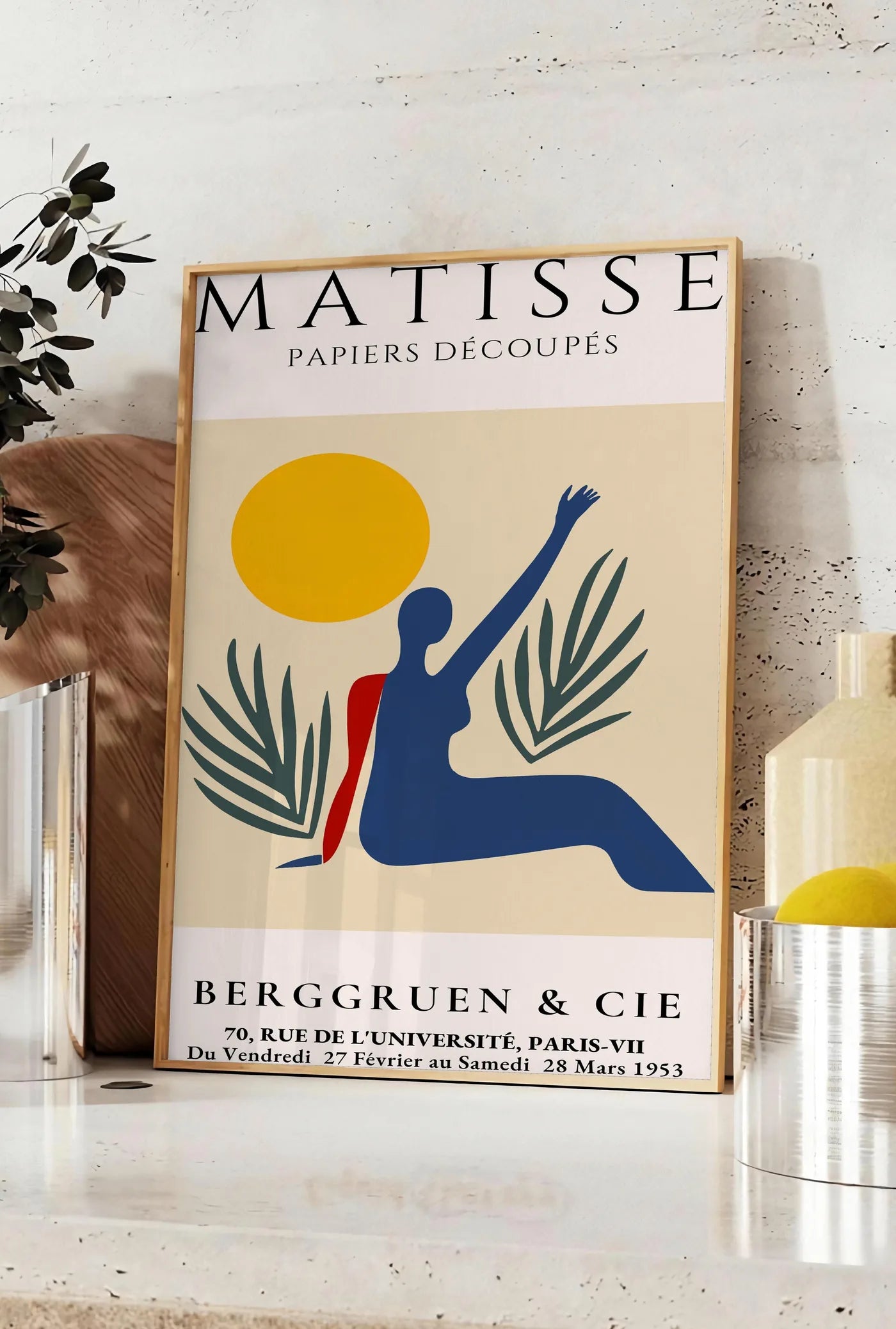 Affiche femme bleue au bras leve sous un soleil jaune et des palmes, posee sur un plan de travail de cuisine en pierre claire, entre une branche d'eucalyptus en vase et un bocal de citrons