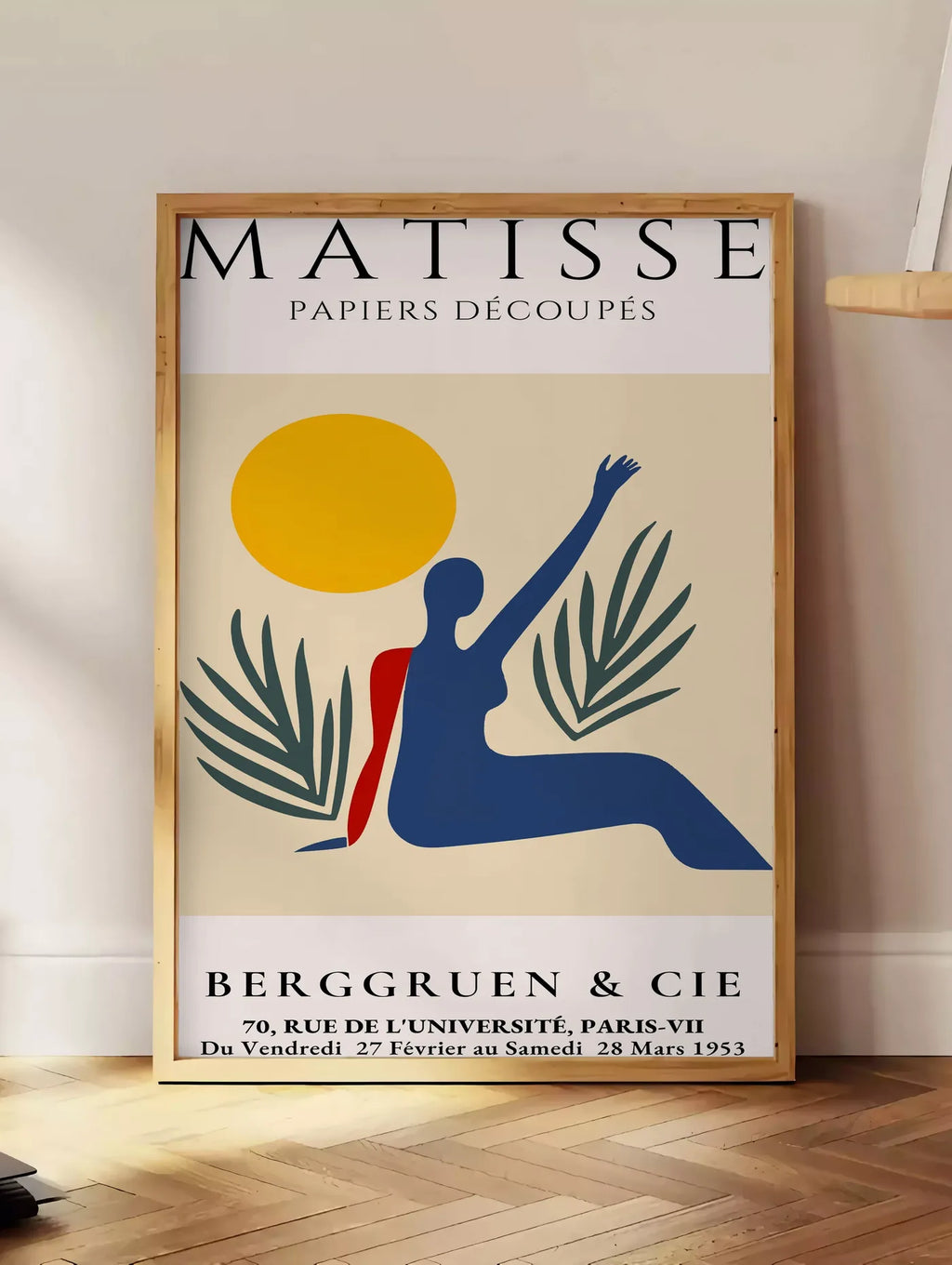 Estampe femme bleue au bras leve sous un soleil jaune et des palmes, posee au sol dans un coin salon, sur un parquet a chevrons, pres d'une table d'appoint en chene chargee de livres