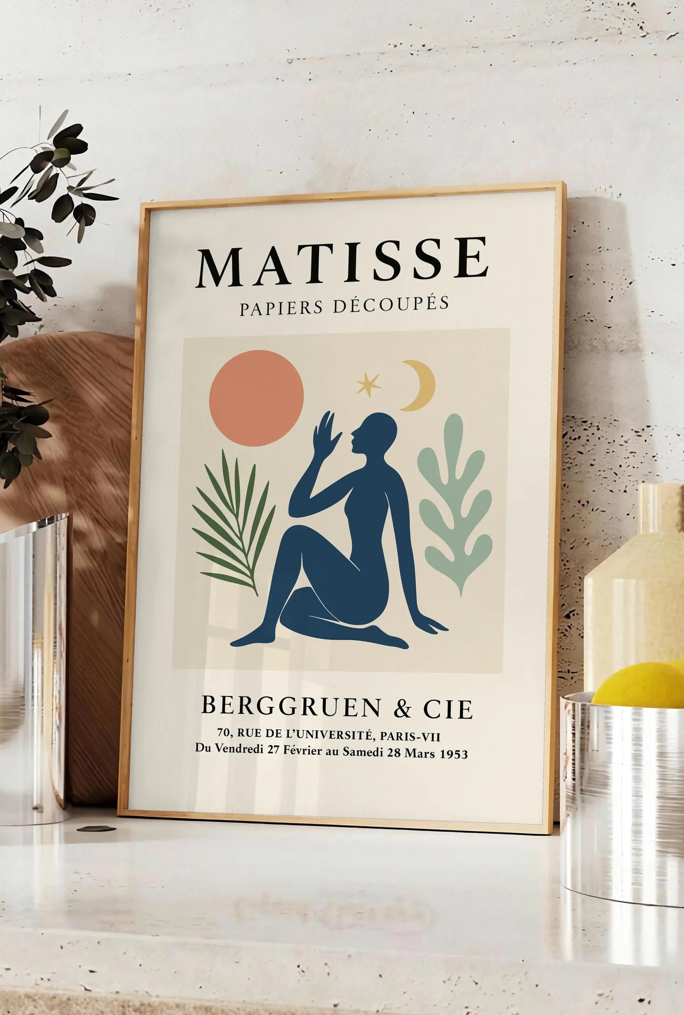 Affiche femme bleue avec lune croissant et etoile, posee sur un plan de travail de cuisine en pierre claire, entre une branche d'eucalyptus en vase et un bocal de citrons