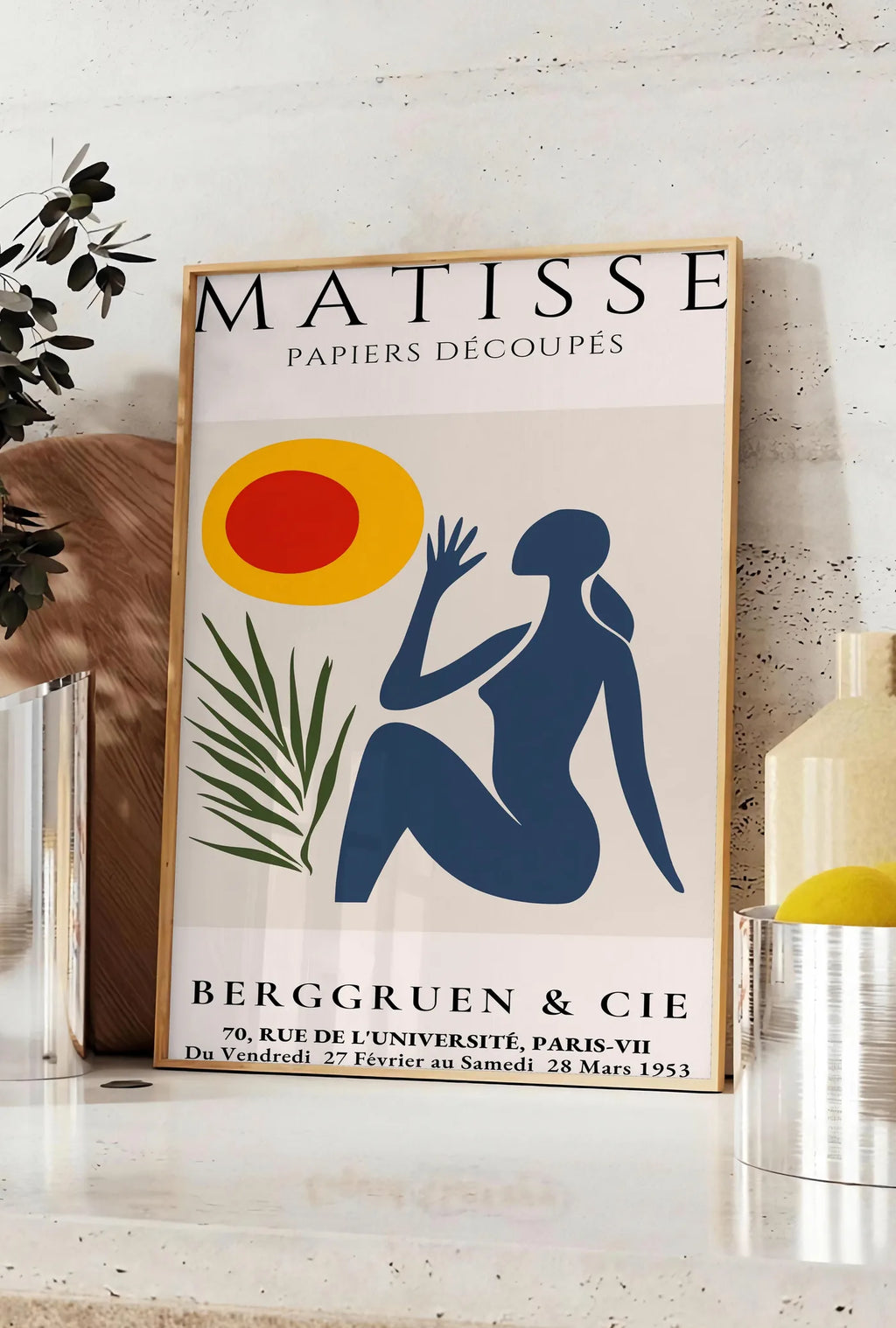 Affiche femme bleue tendue vers un soleil rouge et jaune et une palme, posee sur un plan de travail de cuisine en pierre claire, entre une branche d'eucalyptus en vase et un bocal de citrons