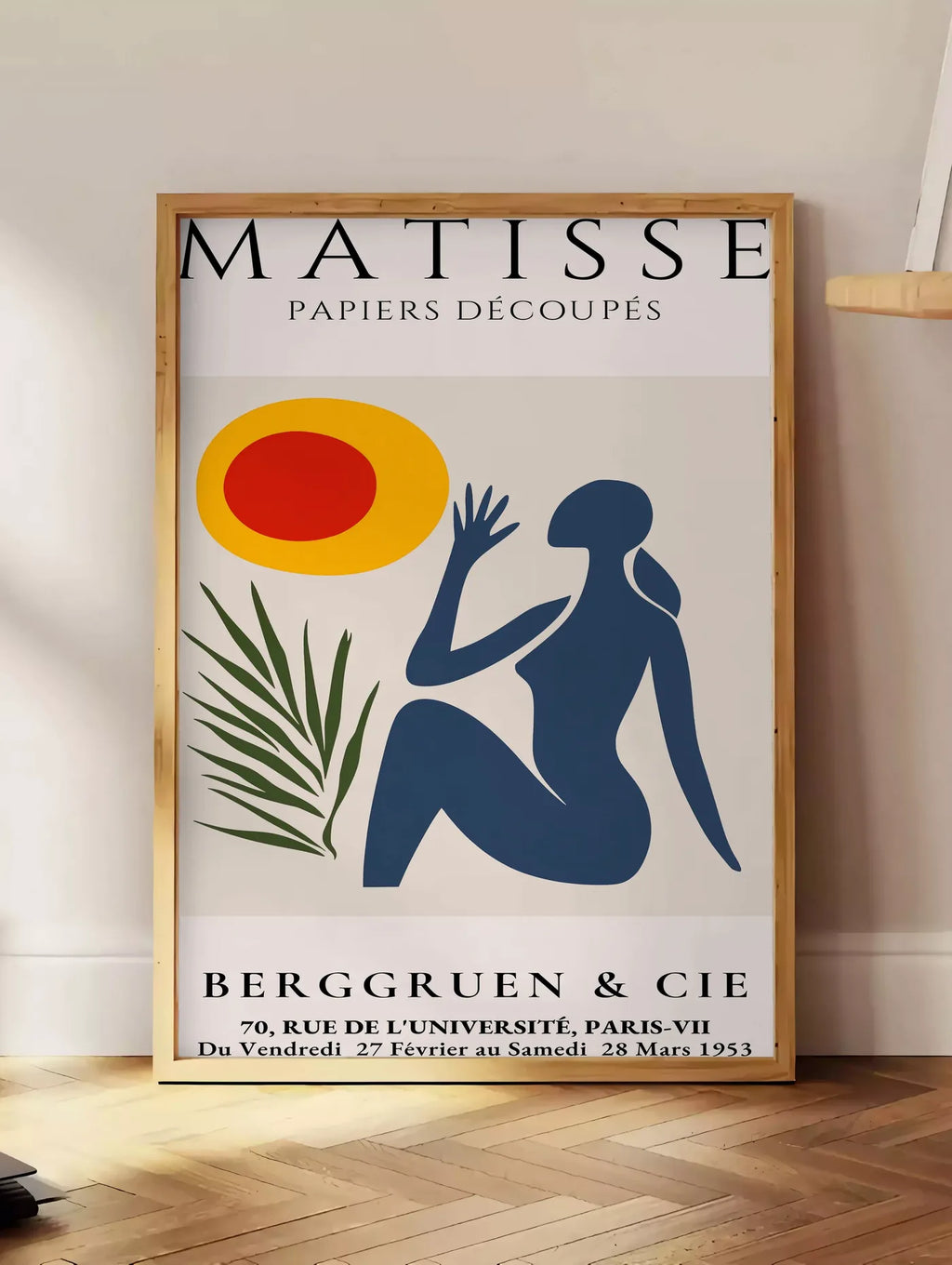 Estampe femme bleue tendue vers un soleil rouge et jaune et une palme, posee au sol dans un coin salon, sur un parquet a chevrons, pres d'une table d'appoint en chene chargee de livres