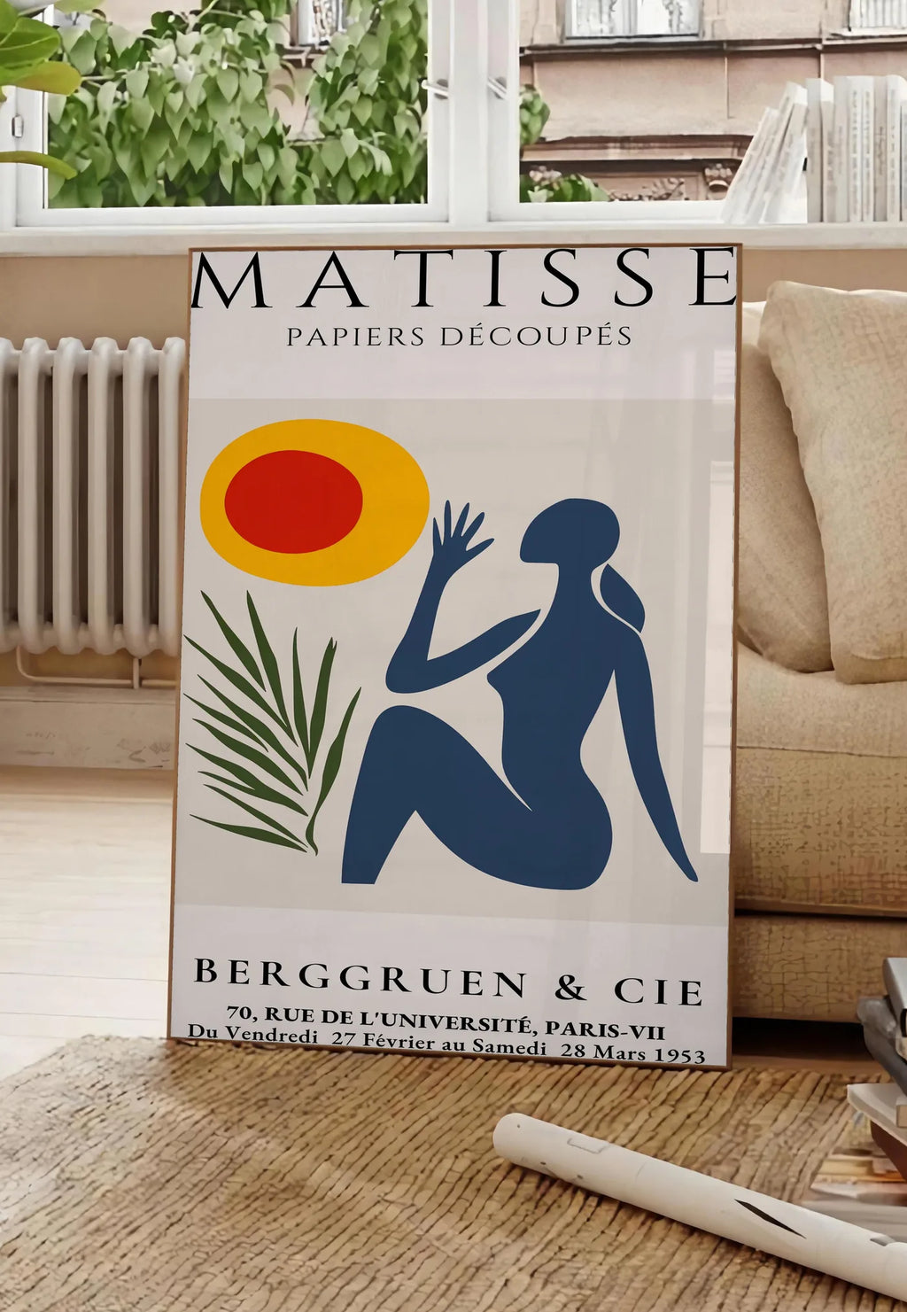 Poster femme bleue tendue vers un soleil rouge et jaune et une palme, posee au sol dans un salon parisien, devant une grande fenetre donnant sur un immeuble haussmannien, entre un ficus et un canape creme