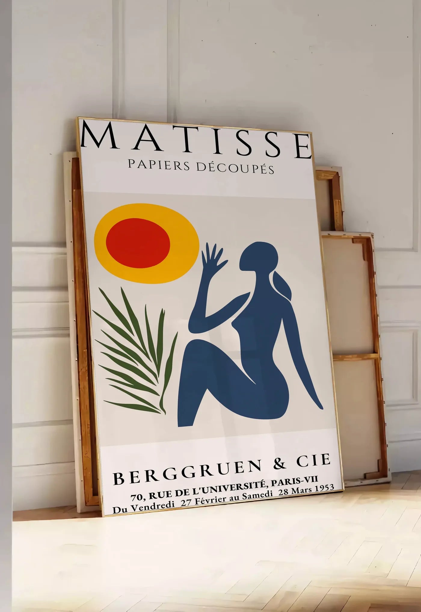 Tirage encadrable femme bleue tendue vers un soleil rouge et jaune et une palme, posee au sol dans une entree lumineuse, contre un mur a panneaux blancs, a cote de chassis en bois empiles