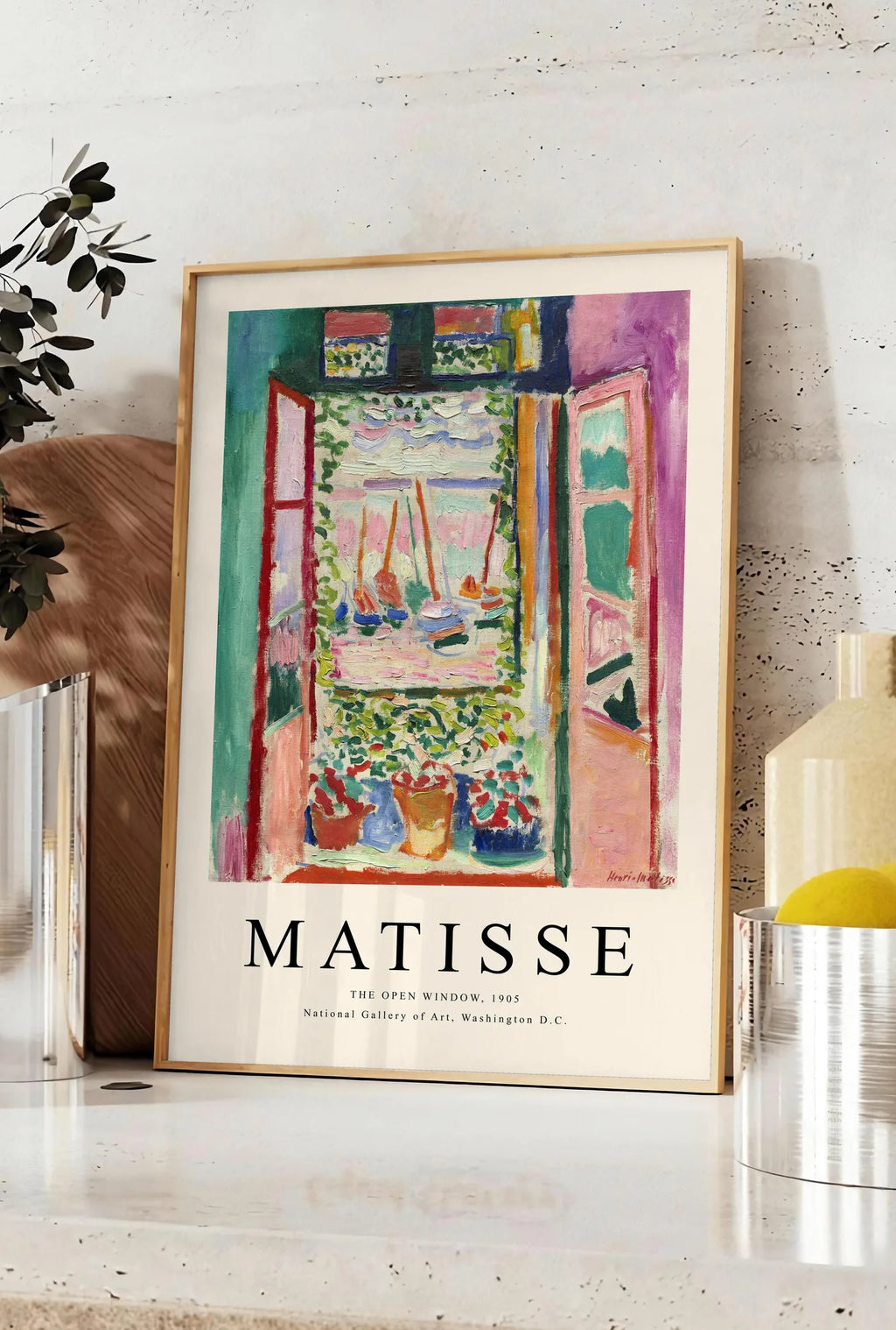 Affiche fenetre ouverte sur une mer rose et turquoise aux voiliers, posee sur un plan de travail de cuisine en pierre claire, entre une branche d'eucalyptus en vase et un bocal de citrons