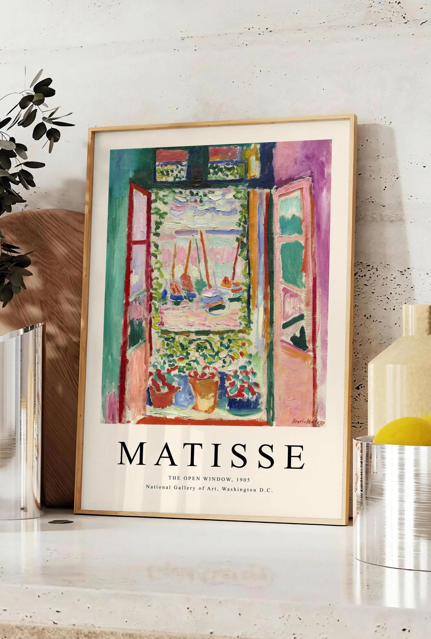 Affiche fenetre ouverte sur une mer rose et turquoise aux voiliers, posee sur un plan de travail de cuisine en pierre claire, entre une branche d'eucalyptus en vase et un bocal de citrons