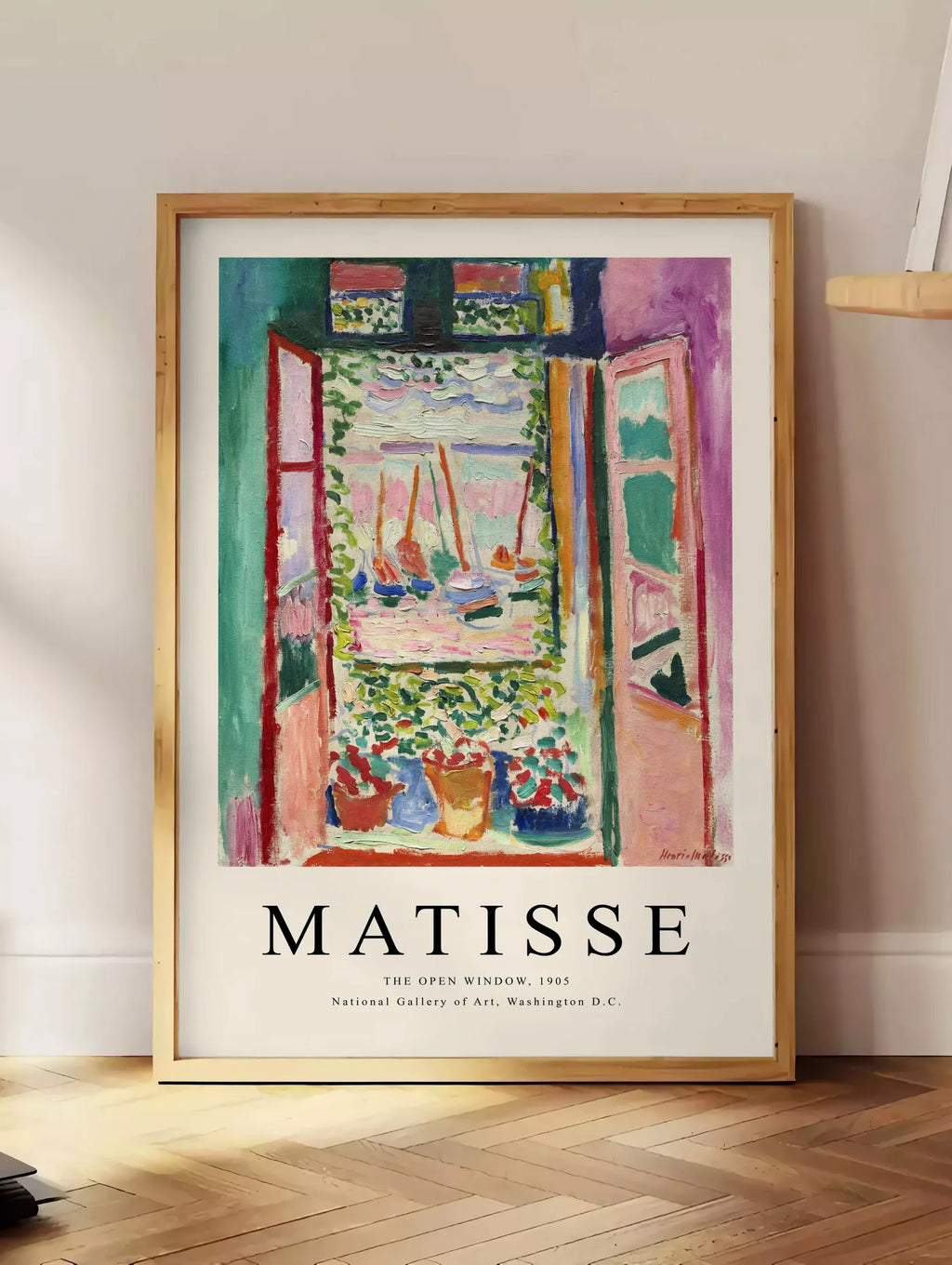 Estampe fenetre ouverte sur une mer rose et turquoise aux voiliers, posee au sol dans un coin salon, sur un parquet a chevrons, pres d'une table d'appoint en chene chargee de livres
