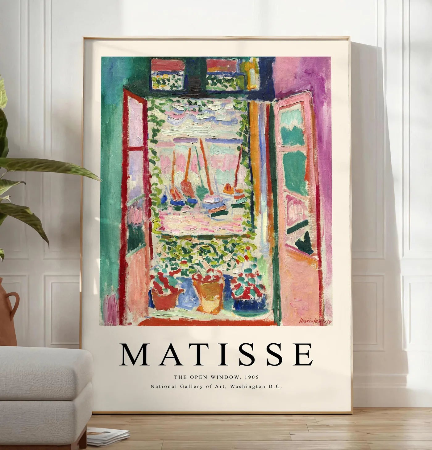 Reproduction fenetre ouverte sur une mer rose et turquoise aux voiliers, posee au sol dans un salon minimaliste clair, a cote d'un canape beige et d'une plante verte, sur un parquet clair