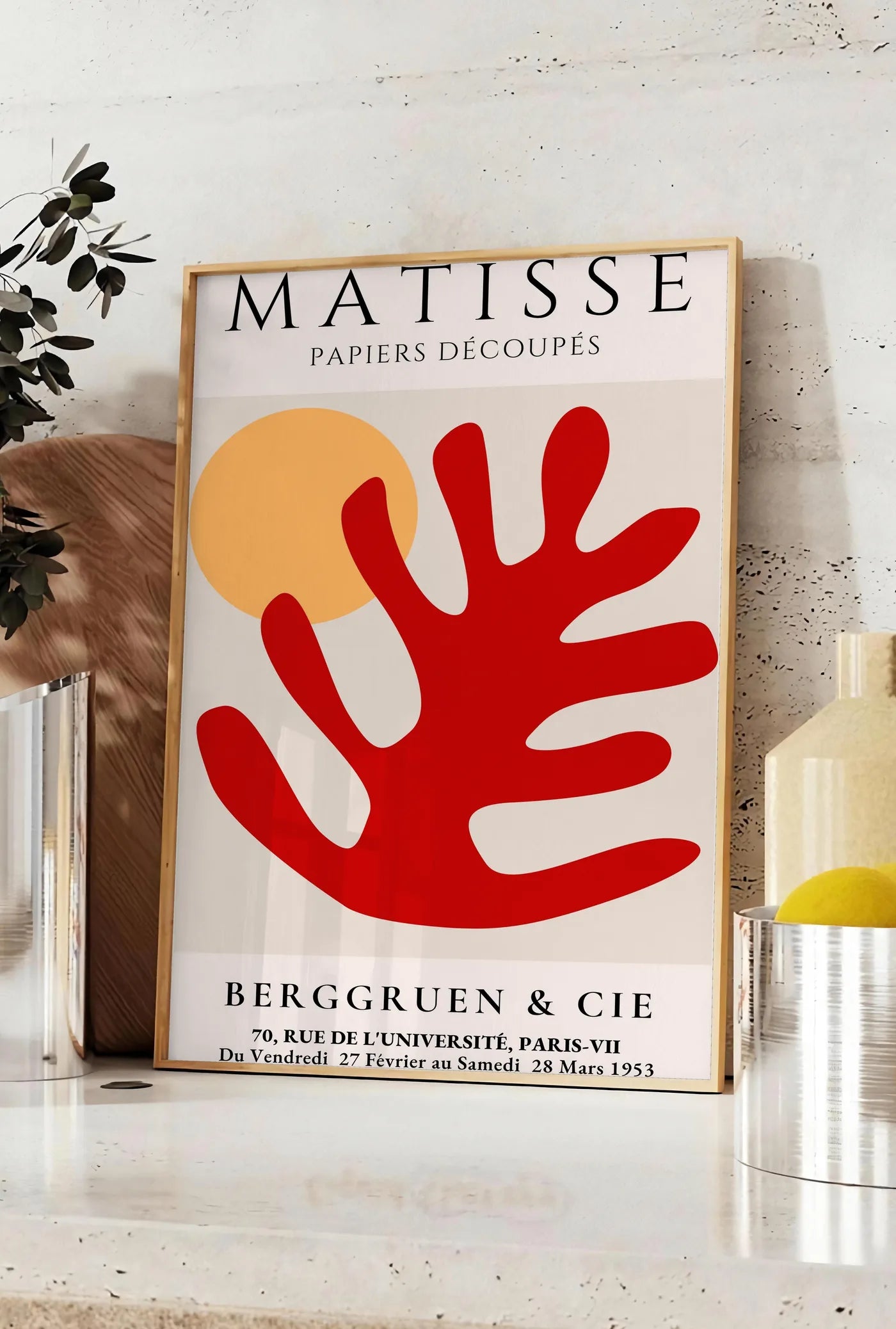 Affiche feuille corail rouge et soleil orange sur fond creme, posee sur un plan de travail de cuisine en pierre claire, entre une branche d'eucalyptus en vase et un bocal de citrons