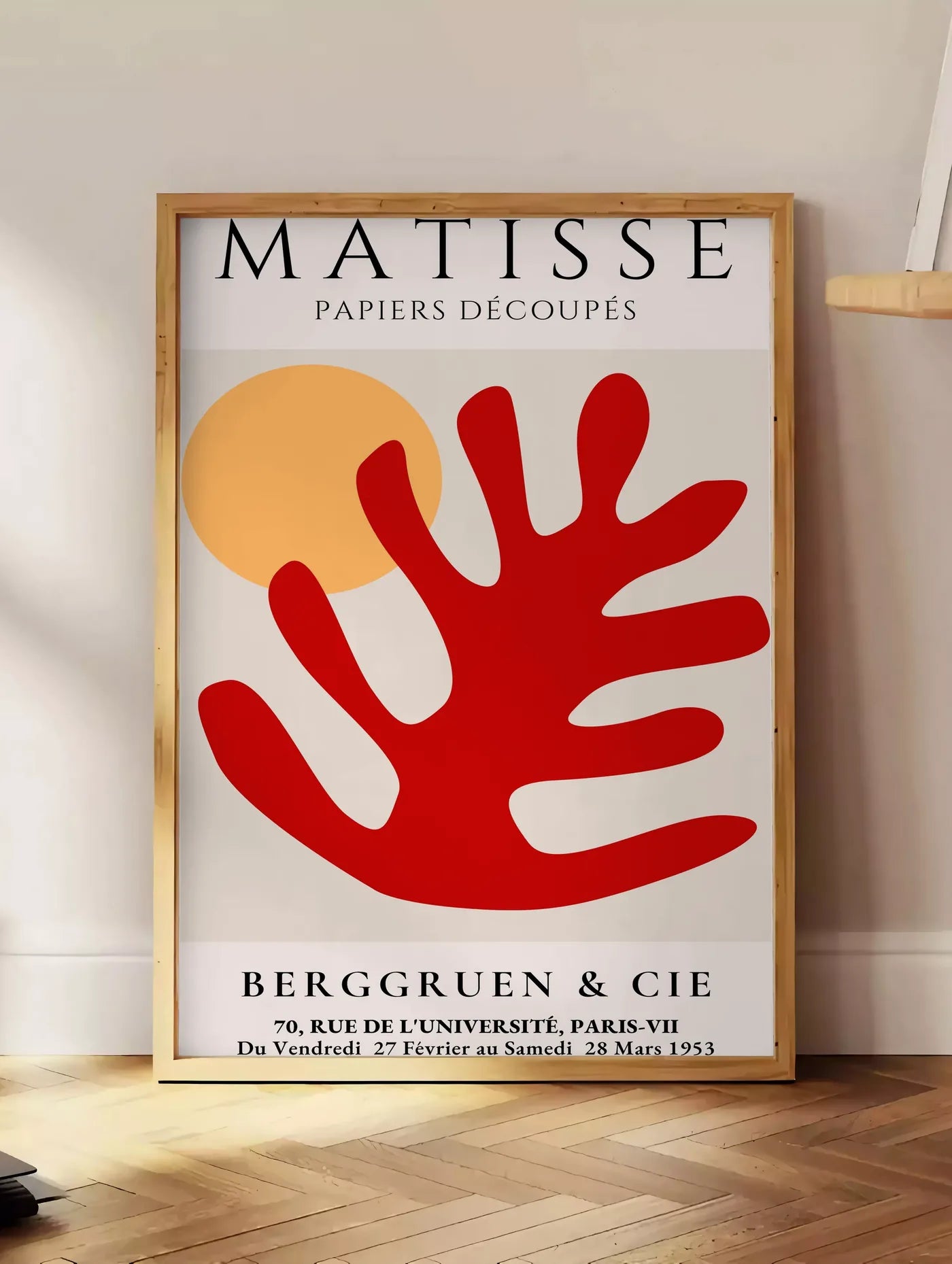 Estampe feuille corail rouge et soleil orange sur fond creme, posee au sol dans un coin salon, sur un parquet a chevrons, pres d'une table d'appoint en chene chargee de livres