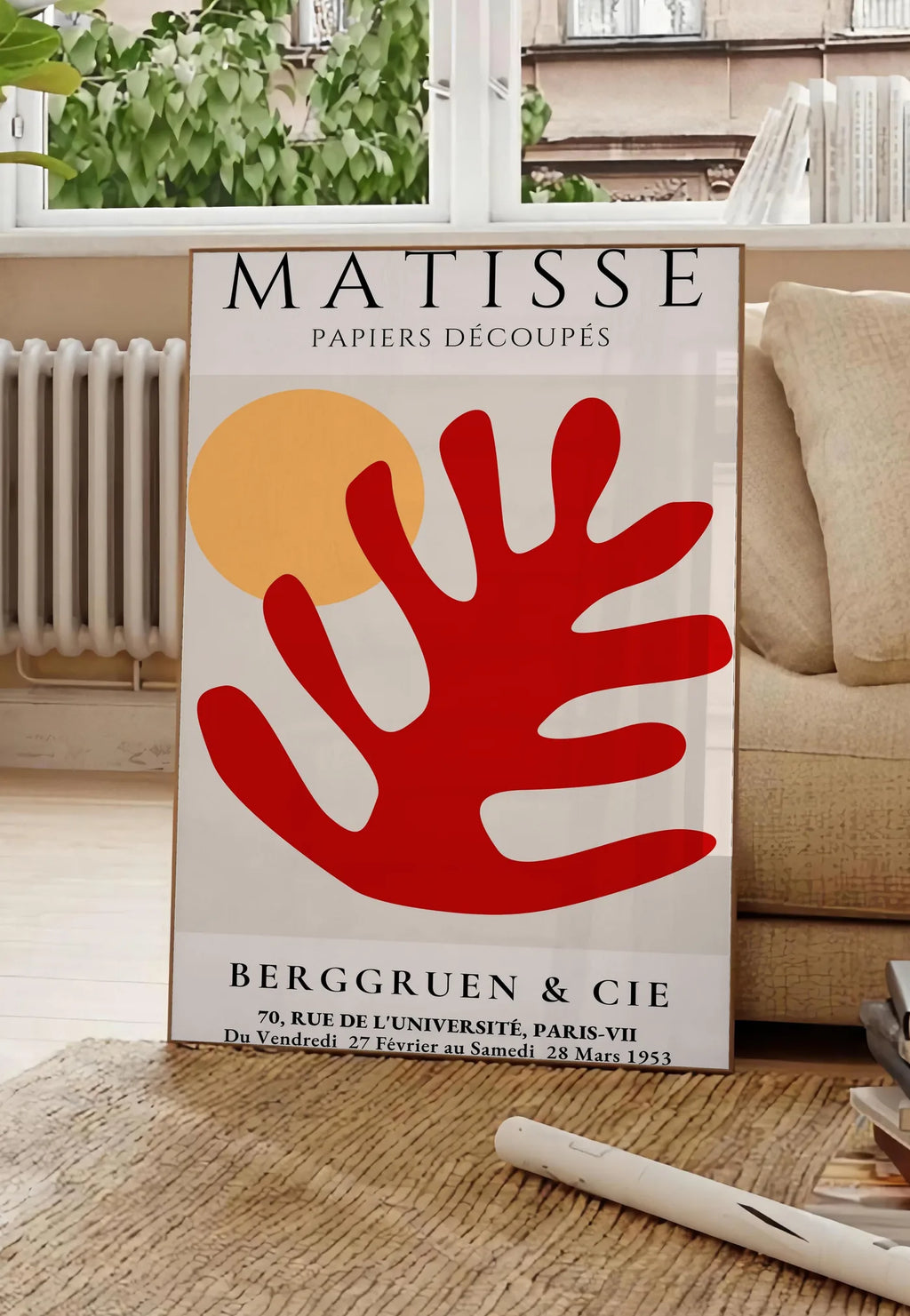 Poster feuille corail rouge et soleil orange sur fond creme, posee au sol dans un salon parisien, devant une grande fenetre donnant sur un immeuble haussmannien, entre un ficus et un canape creme