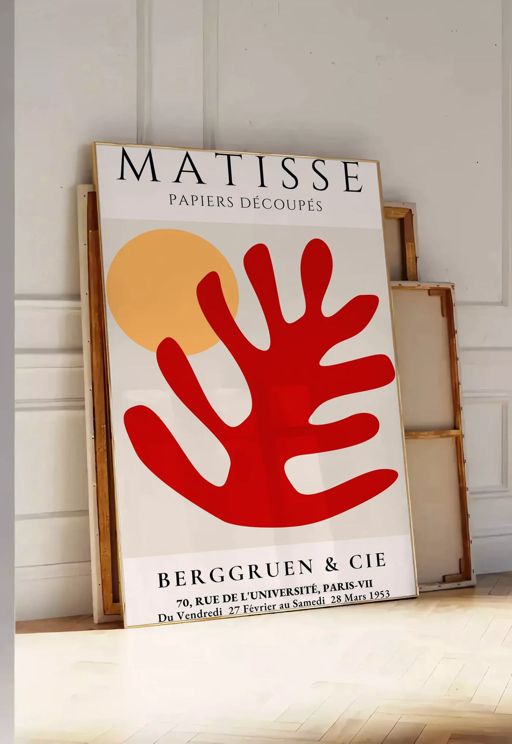 Tirage encadrable feuille corail rouge et soleil orange sur fond creme, posee au sol dans une entree lumineuse, contre un mur a panneaux blancs, a cote de chassis en bois empiles