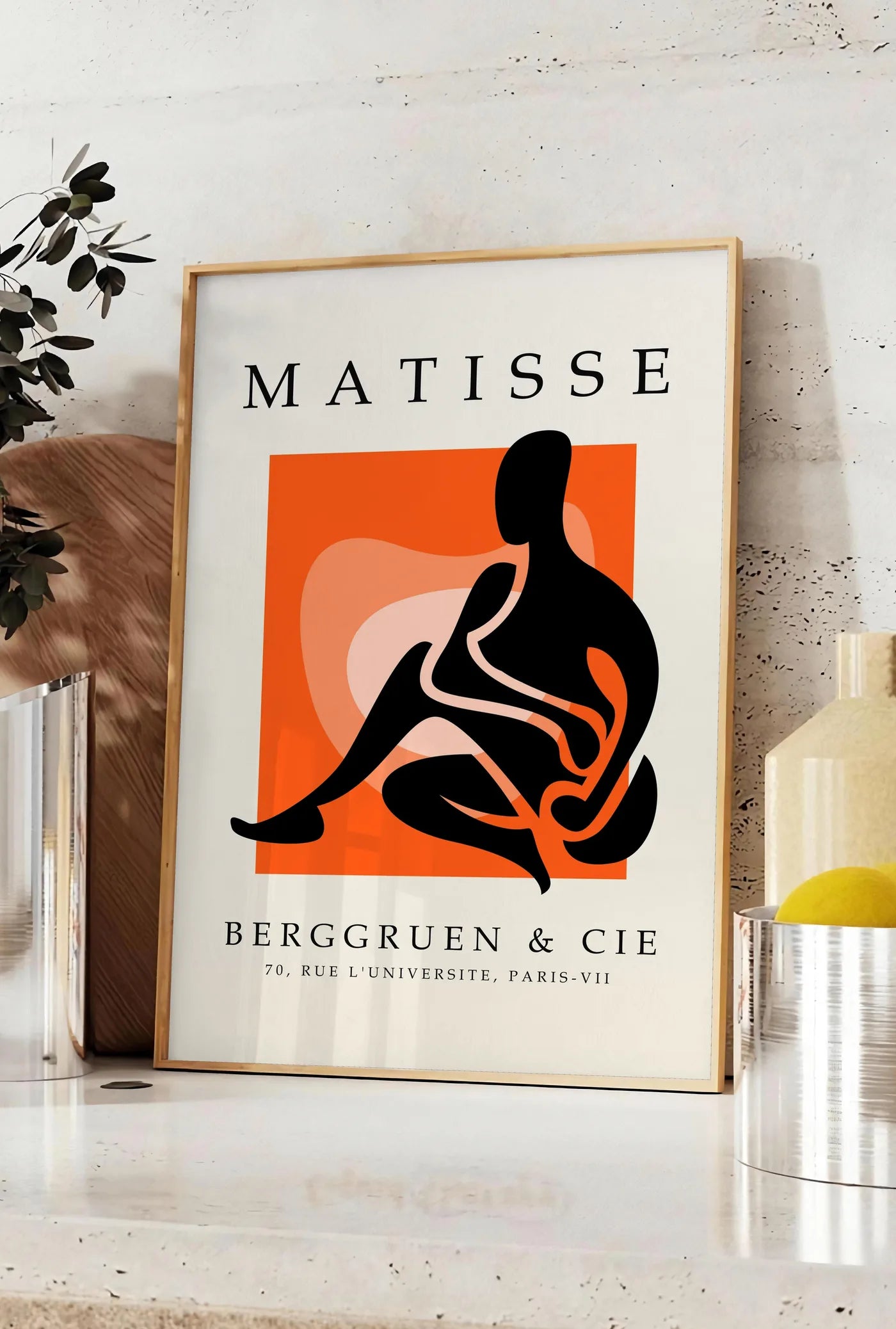 Affiche figure noire au trait blanc sur carre orange, posee sur un plan de travail de cuisine en pierre claire, entre une branche d'eucalyptus en vase et un bocal de citrons