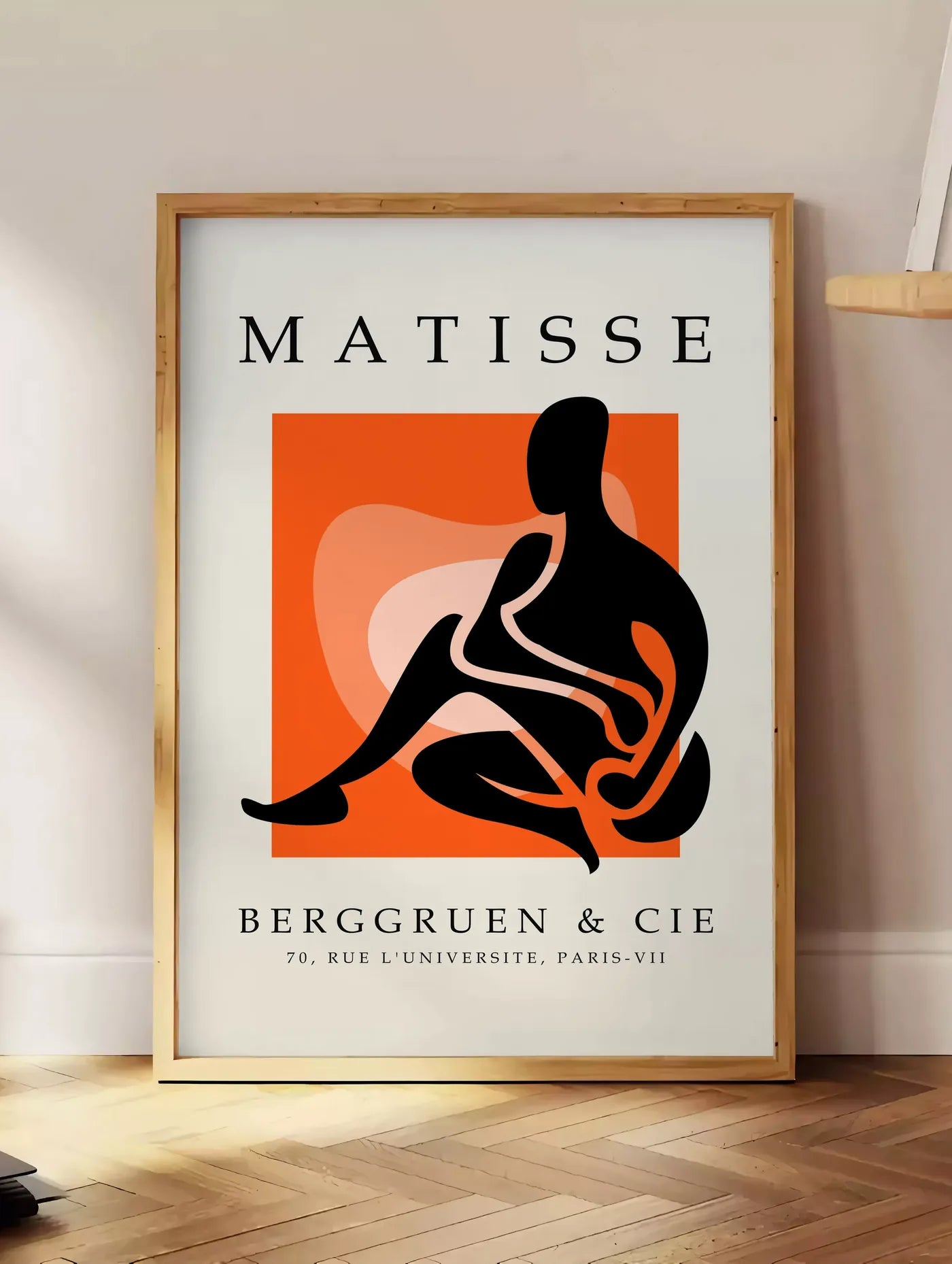 Estampe figure noire au trait blanc sur carre orange, posee au sol dans un coin salon, sur un parquet a chevrons, pres d'une table d'appoint en chene chargee de livres