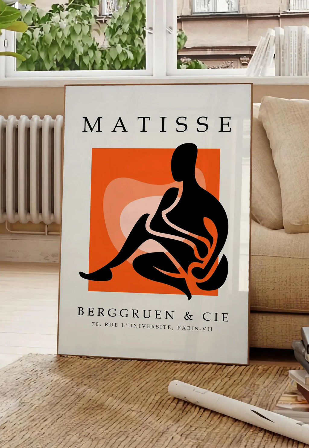 Poster figure noire au trait blanc sur carre orange, posee au sol dans un salon parisien, devant une grande fenetre donnant sur un immeuble haussmannien, entre un ficus et un canape creme