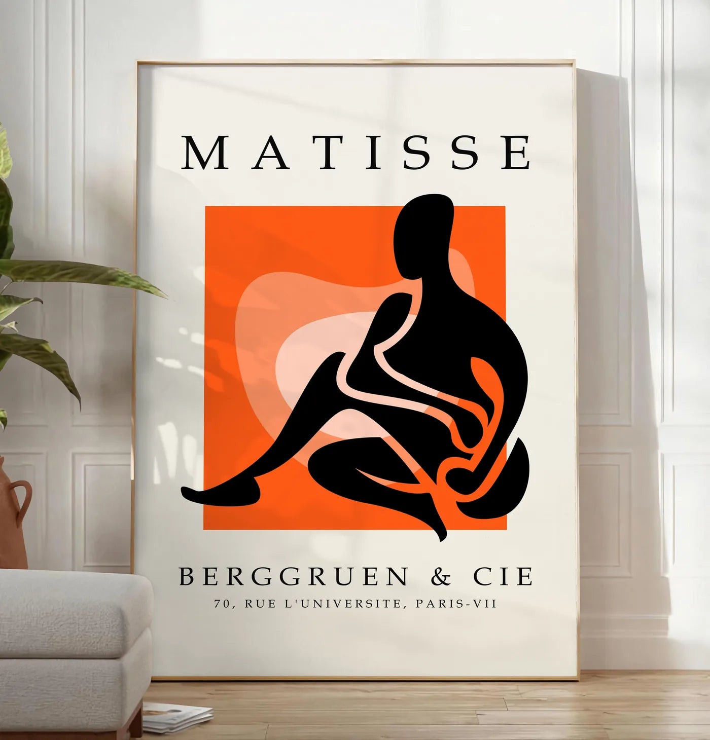 Reproduction figure noire au trait blanc sur carre orange, posee au sol dans un salon minimaliste clair, a cote d'un canape beige et d'une plante verte, sur un parquet clair
