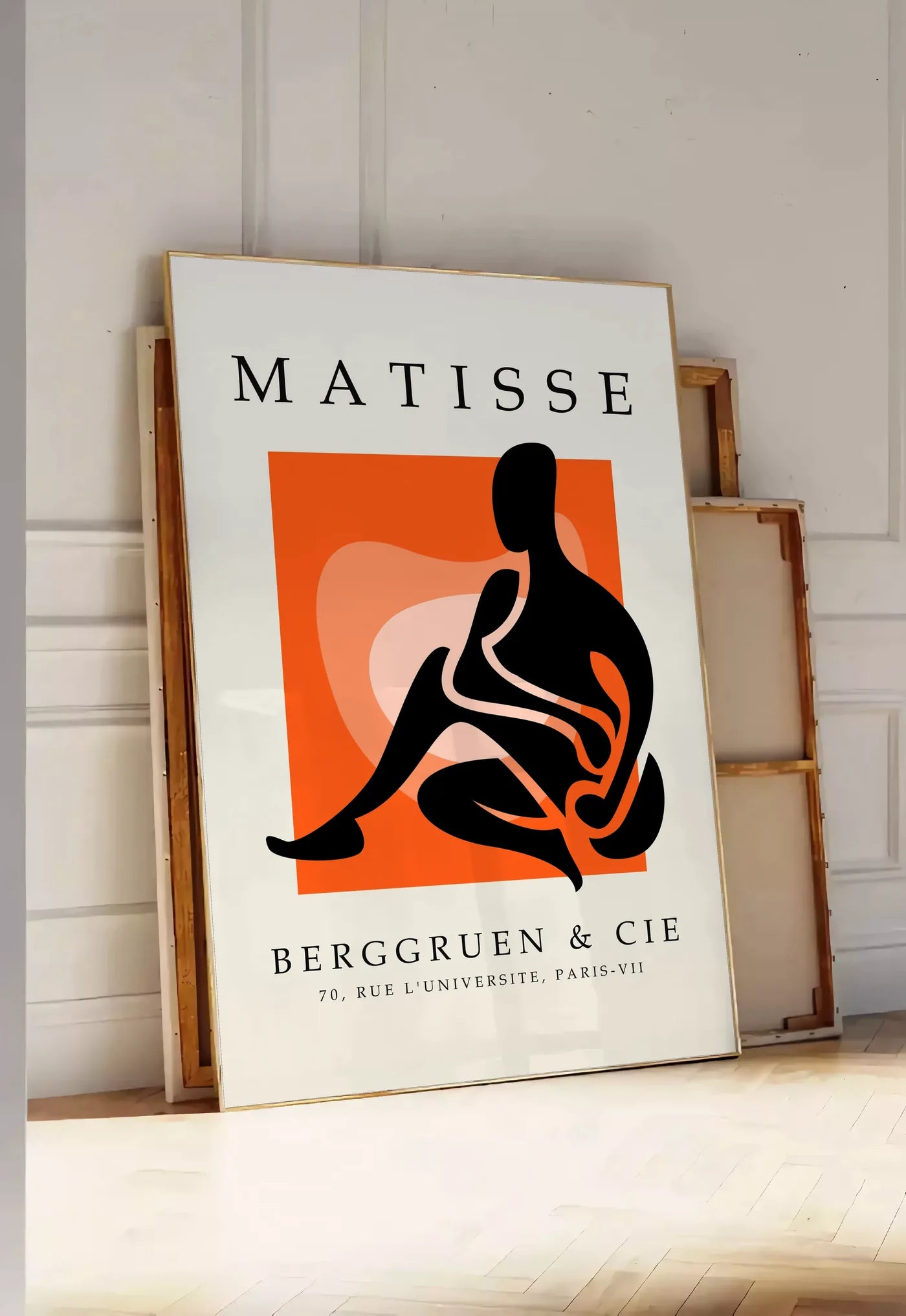 Tirage encadrable figure noire au trait blanc sur carre orange, posee au sol dans une entree lumineuse, contre un mur a panneaux blancs, a cote de chassis en bois empiles