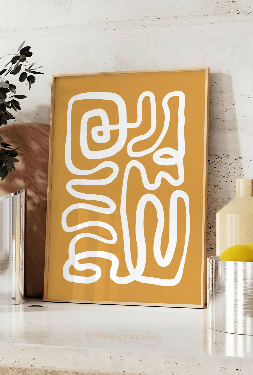 Affiche ligne continue blanche abstraite sur fond ocre, posee sur un plan de travail de cuisine en pierre claire, entre une branche d'eucalyptus en vase et un bocal de citrons