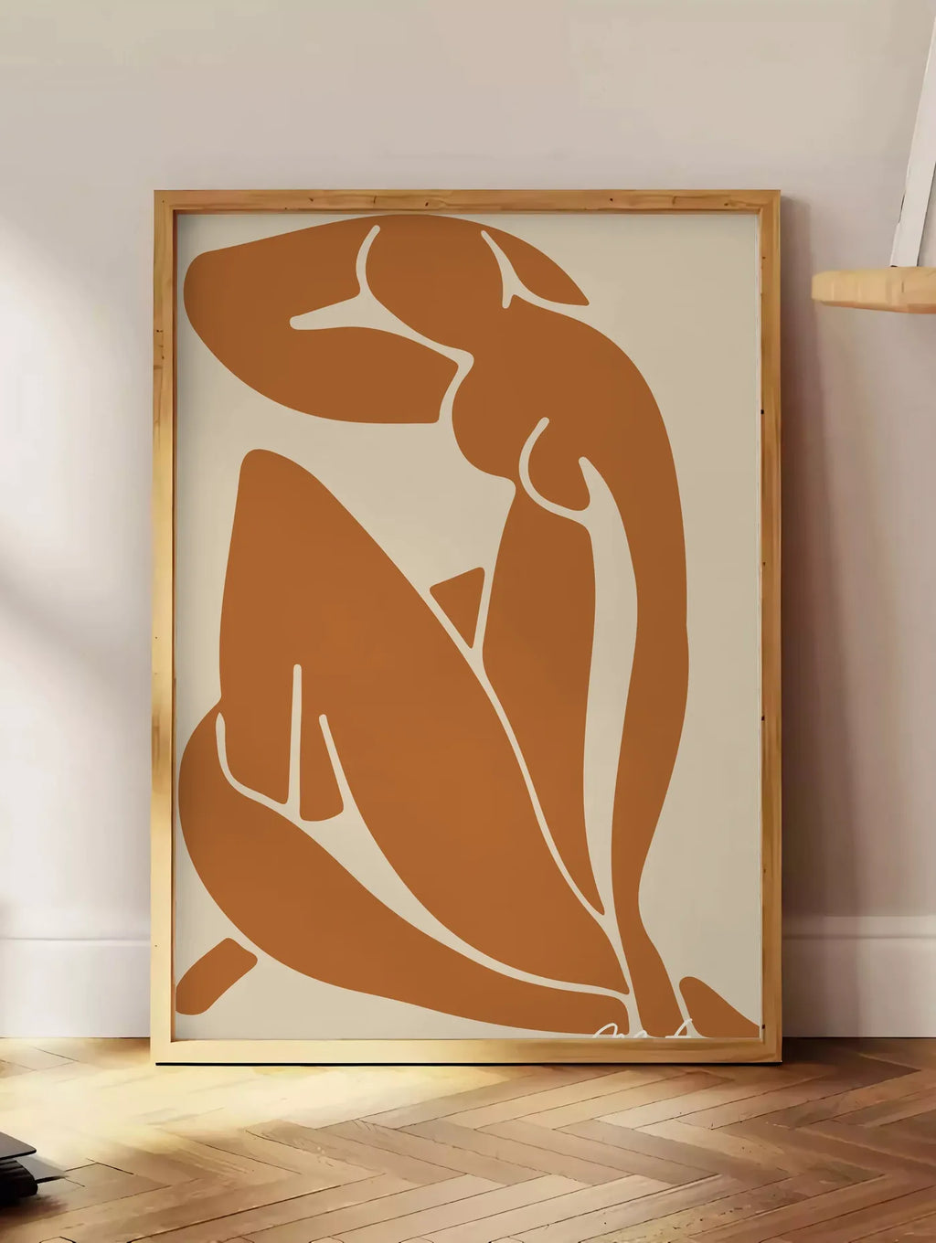 Estampe silhouette nue découpée terracotta sur fond beige, posee au sol dans un coin salon, sur un parquet a chevrons, pres d'une table d'appoint en chene chargee de livres