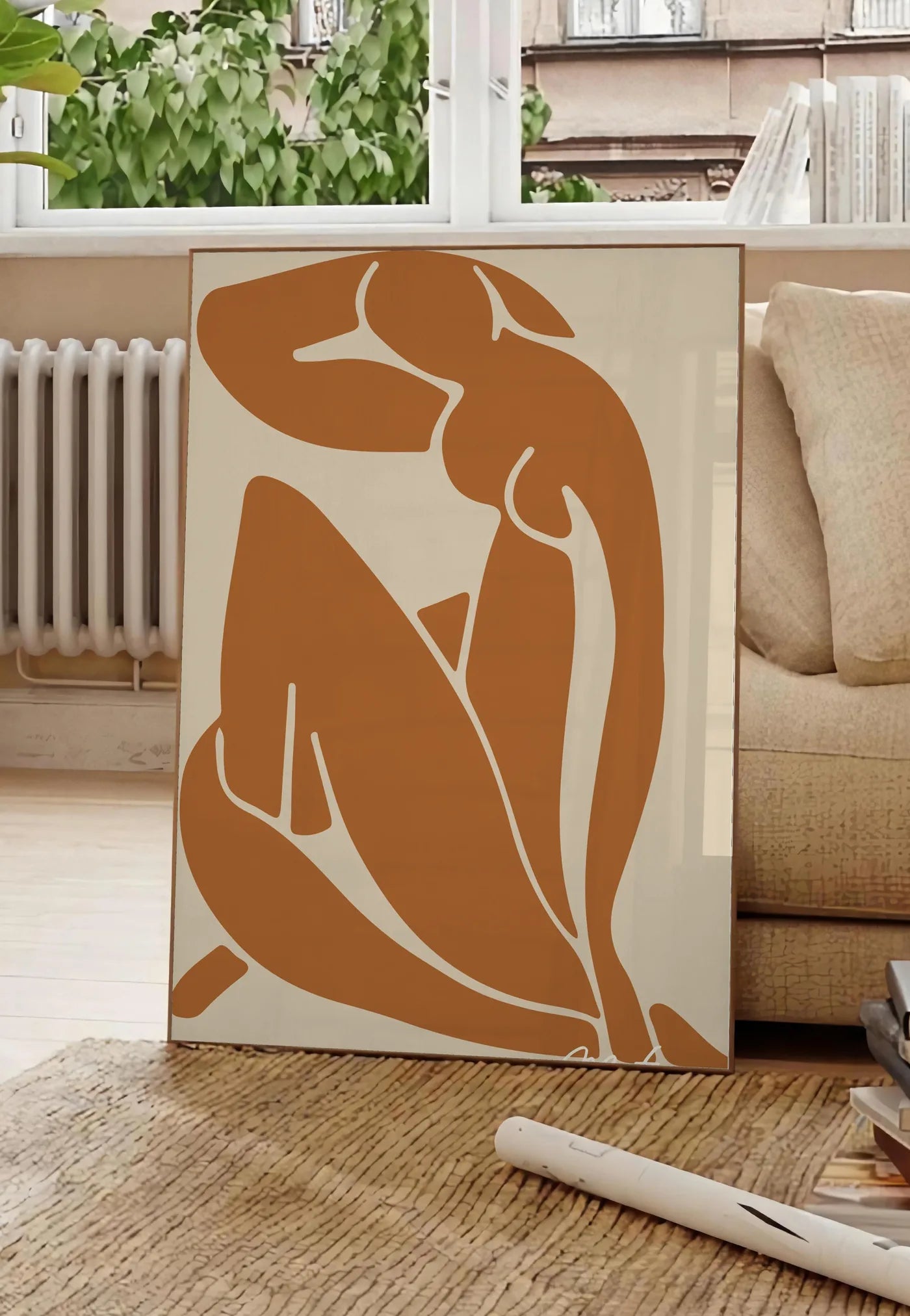 Poster silhouette nue découpée terracotta sur fond beige, posee au sol dans un salon parisien, devant une grande fenetre donnant sur un immeuble haussmannien, entre un ficus et un canape creme