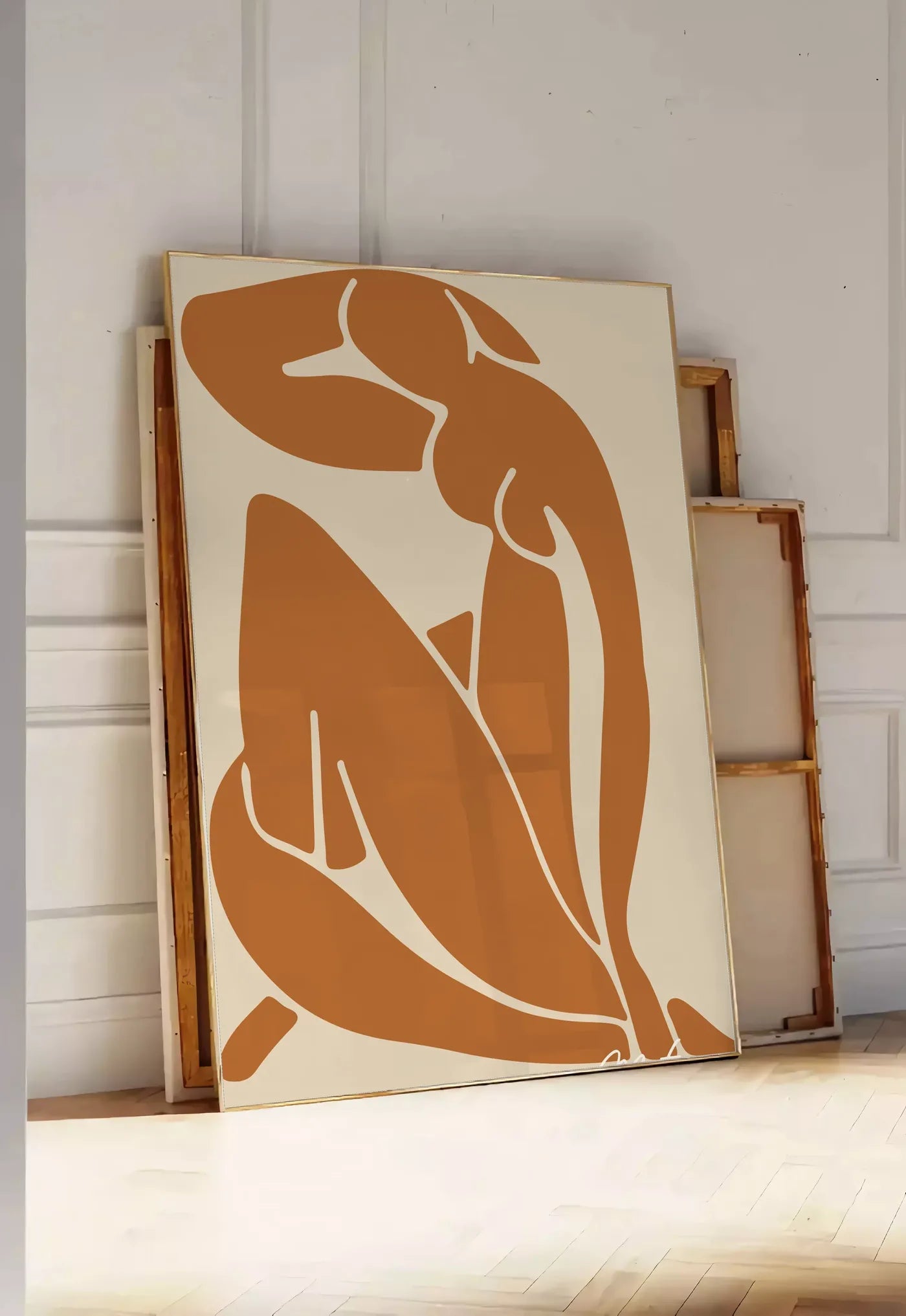 Tirage encadrable silhouette nue découpée terracotta sur fond beige, posee au sol dans une entree lumineuse, contre un mur a panneaux blancs, a cote de chassis en bois empiles