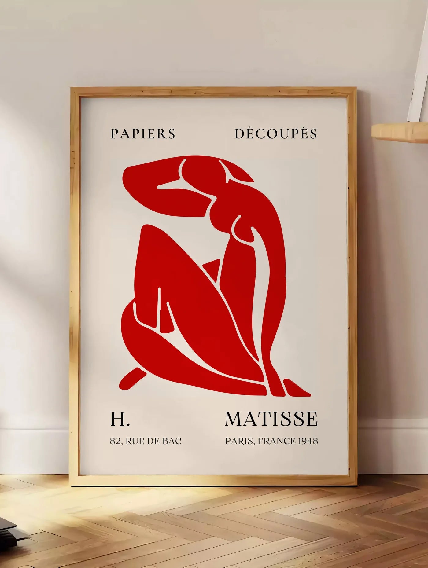 Estampe affiche Matisse Papiers Decoupes, posee au sol dans un coin salon, sur un parquet a chevrons, pres d'une table d'appoint en chene chargee de livres