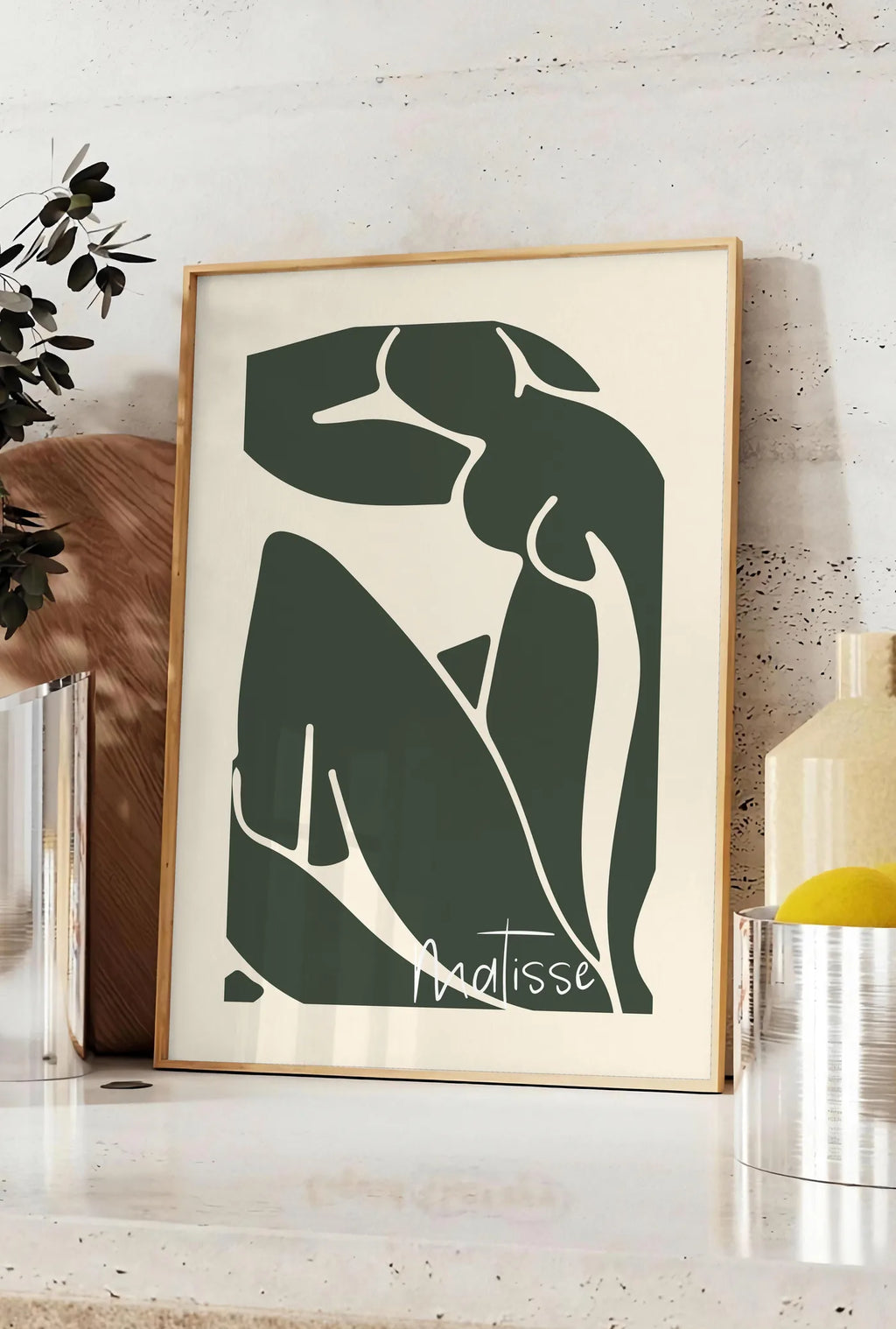 Affiche nu assis vert au trait blanc sur fond creme, posee sur un plan de travail de cuisine en pierre claire, entre une branche d'eucalyptus en vase et un bocal de citrons